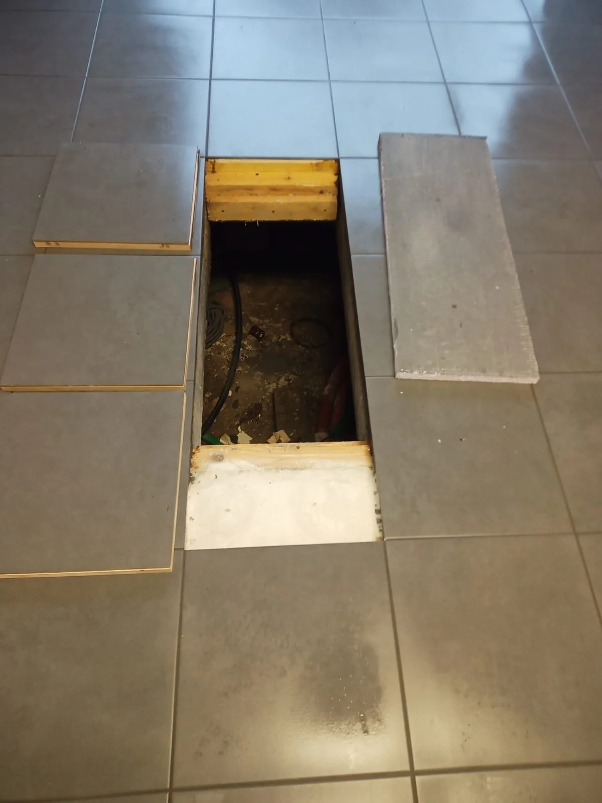 Une trappe de sol ouverte dans un plan de travail de cuisine, révélant un espace de plomberie et d'électricité en dessous, entourée de carreaux de sol gris.