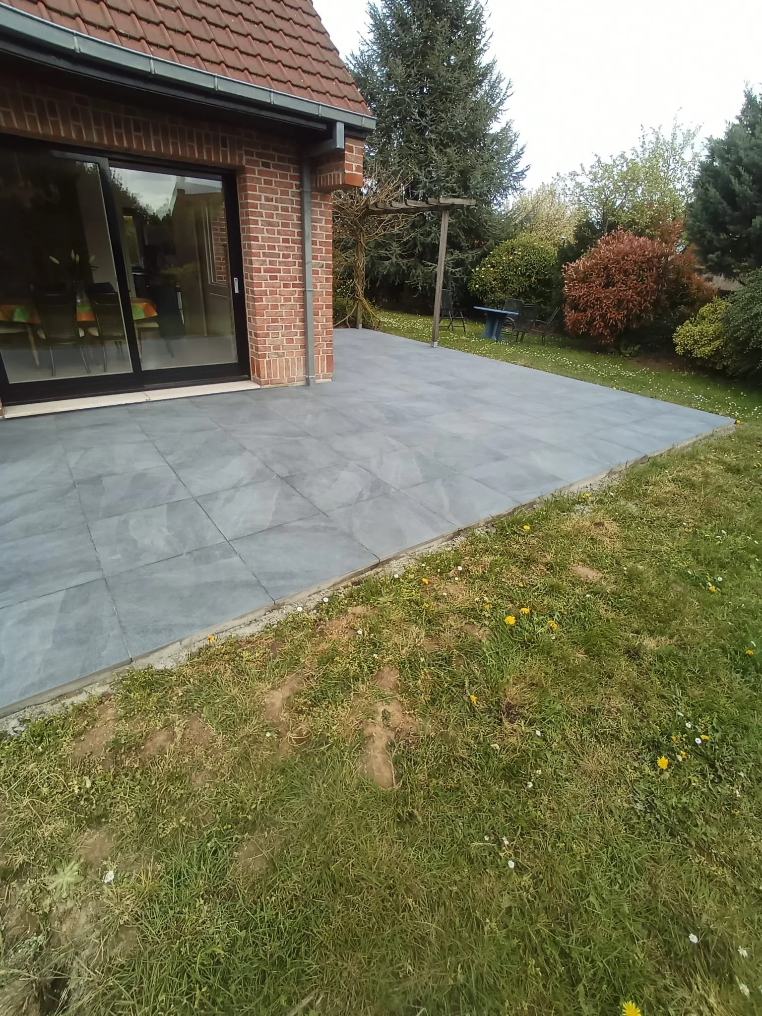 Une terrasse en carrelage gris foncé devant une maison en briques rouges avec une porte-fenêtre, entourée de pelouse verte avec des fleurs, et des arbres en arrière-plan.