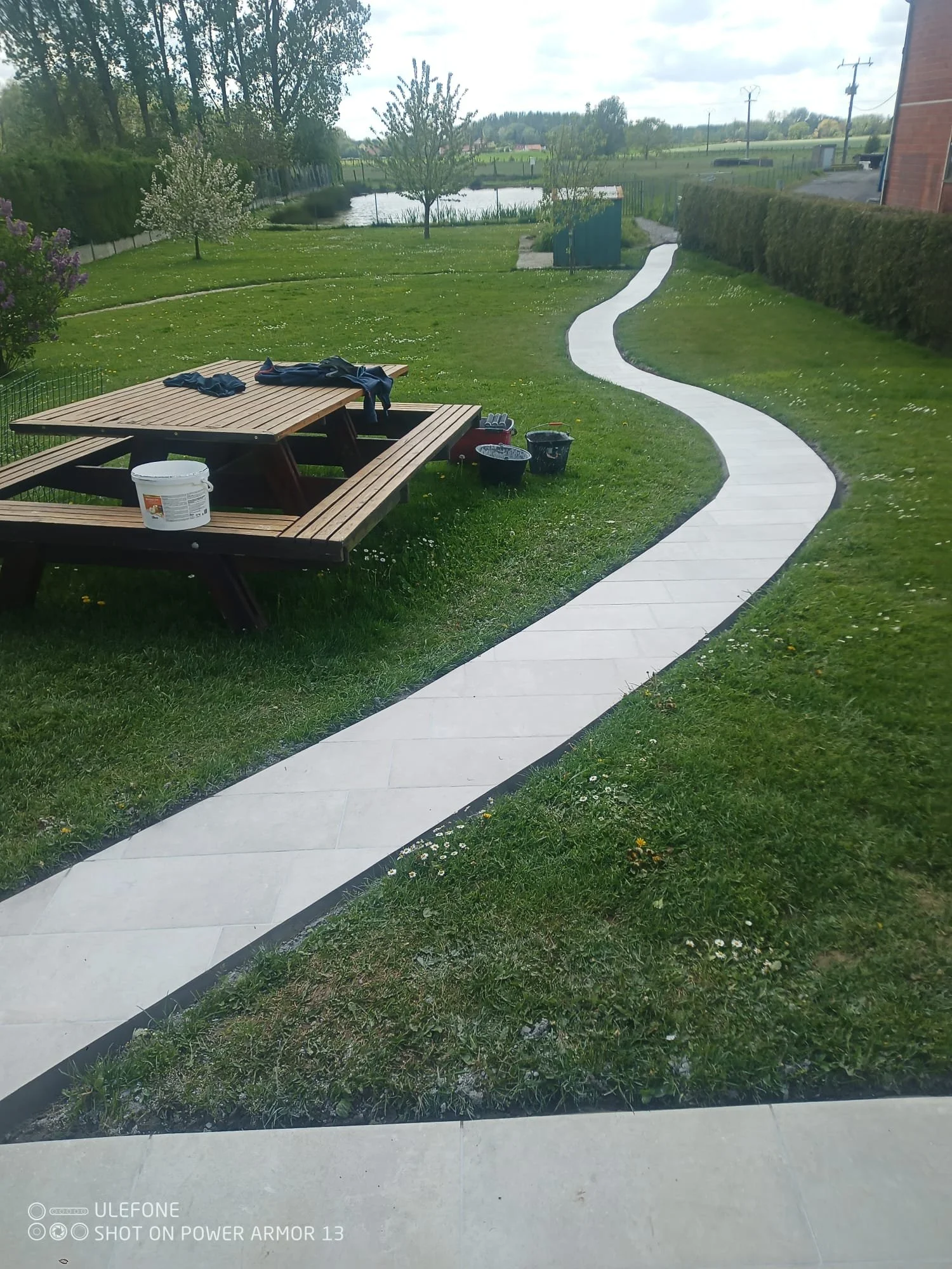 Un jardin bien entretenu avec une pelouse verte, des arbres en fleurs, une petite mare, une allée en pierre blanche serpente à travers la pelouse, et une table en bois avec quelques vêtements et objets posés dessus.