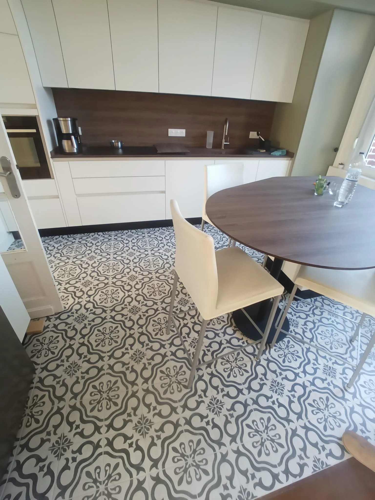 Une cuisine moderne avec un plan de travail en bois foncé, des armoires blanches, un évier, une cafetière, une bouteille d'eau et un verre sur une table ronde en bois. Le sol a un carrelage à motifs noir et blanc.