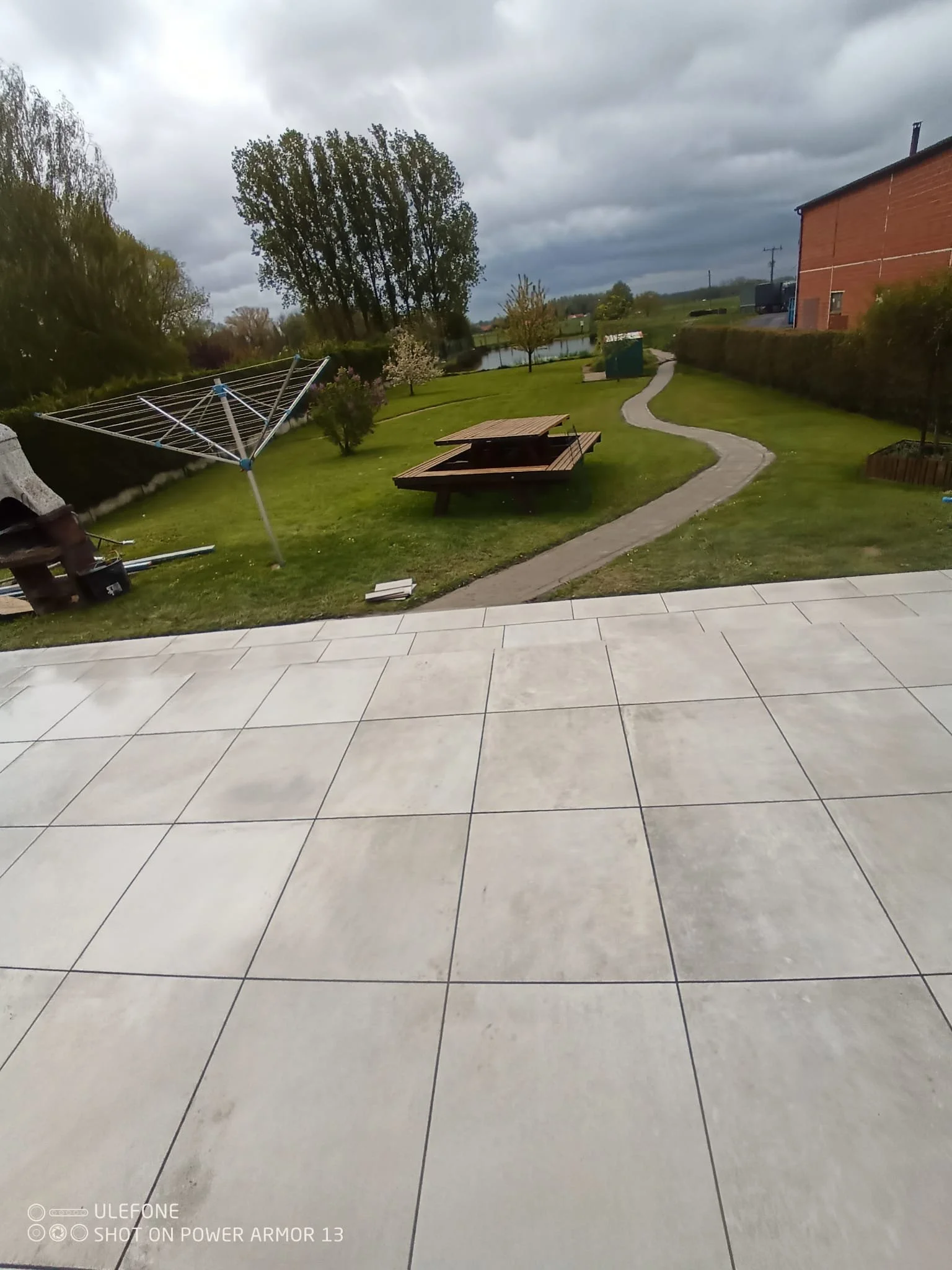 Jardin avec pelouse, allée serpentante, table de pique-nique en bois, arbre, bâtiment, drain de ventilation, étendoir à linge, et un corps d'eau en arrière-plan, sous un ciel nuageux.