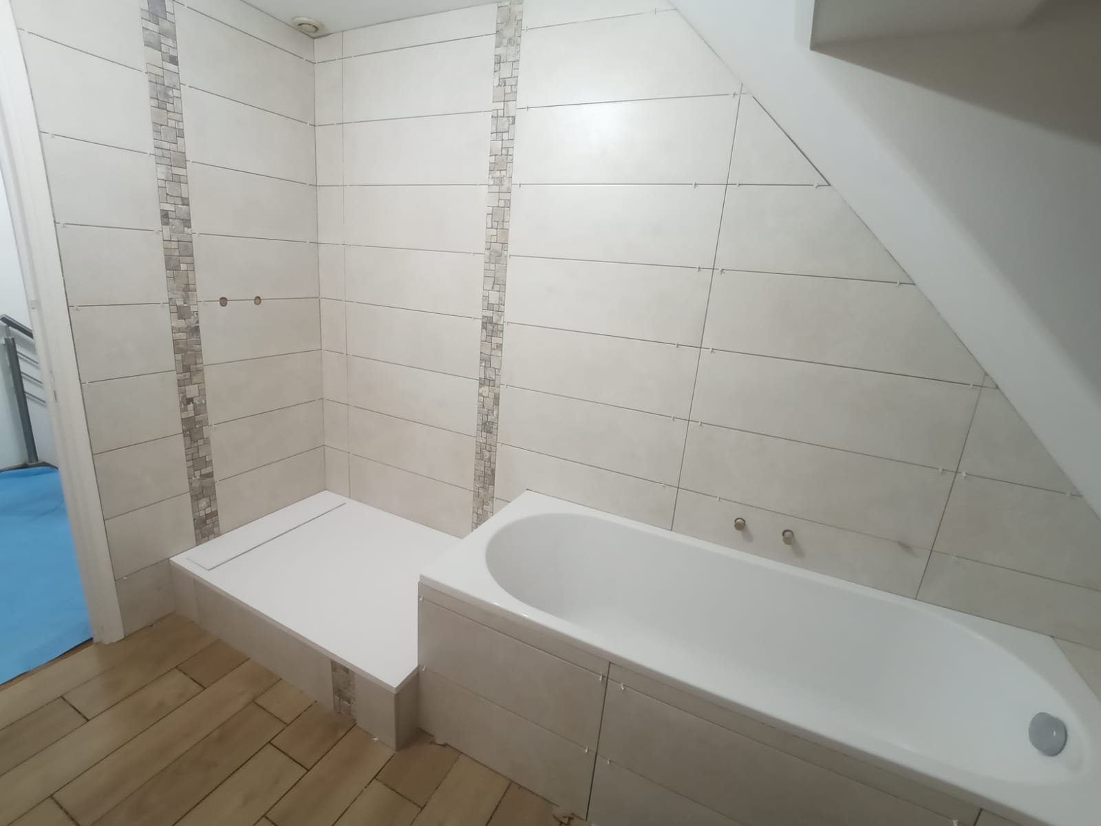 Salle de bain avec une baignoire et un espace de douche, murs en carrelage beige, sol en carrelage imitation bois, mur au-dessus de la baignoire avec une pente de plafond.