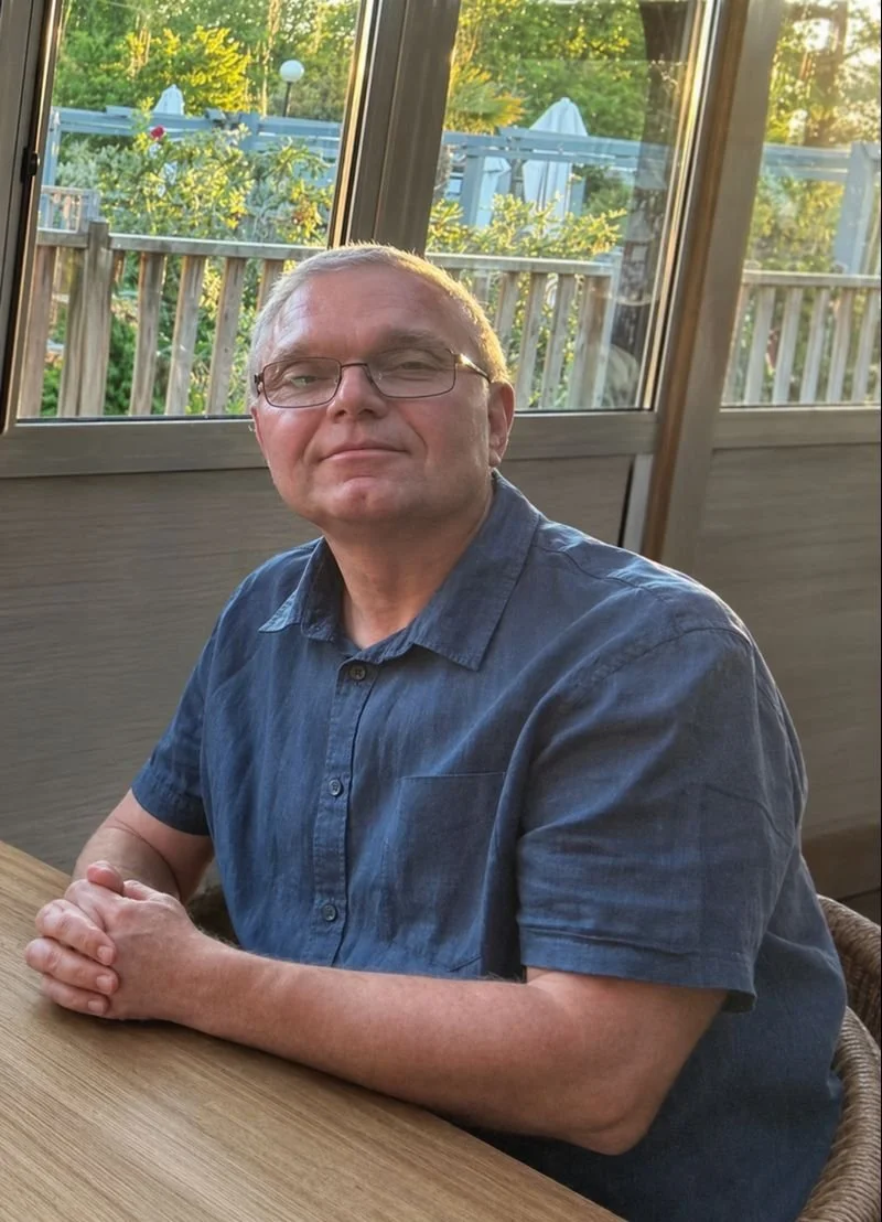 Un homme assis à une table intérieure, portant des lunettes et une chemise en denim, avec une fenêtre derrière lui donnant sur un jardin en plein air avec des parasols blancs.