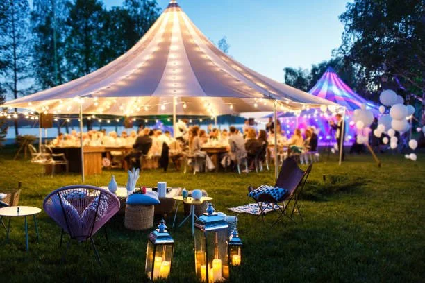 Een feesttent met mensen die aan tafels zitten, omgeven door lampjes en decoraties, met buitenverlichting en gezellige zitplekken op het grasveld.
