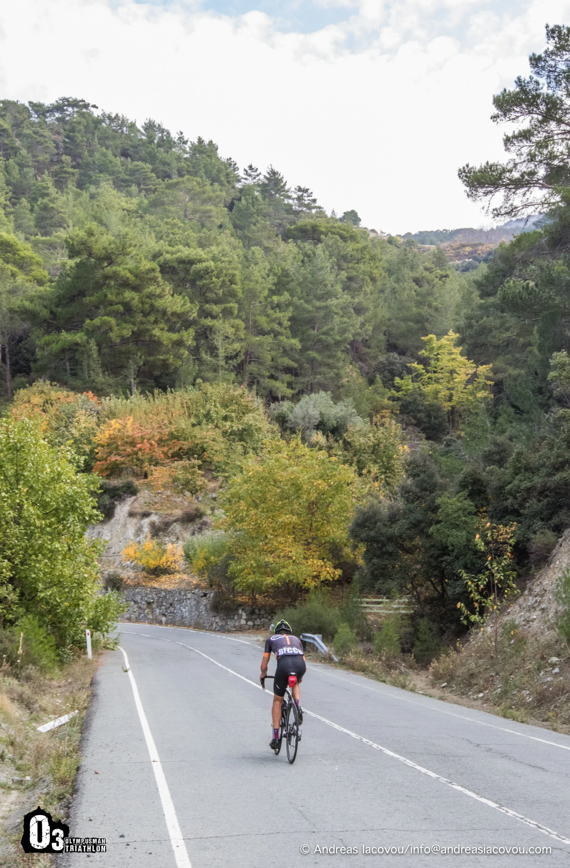 A cyclist riding on a winding mountain road surrounded by lush green trees with a cloudy sky overhead.