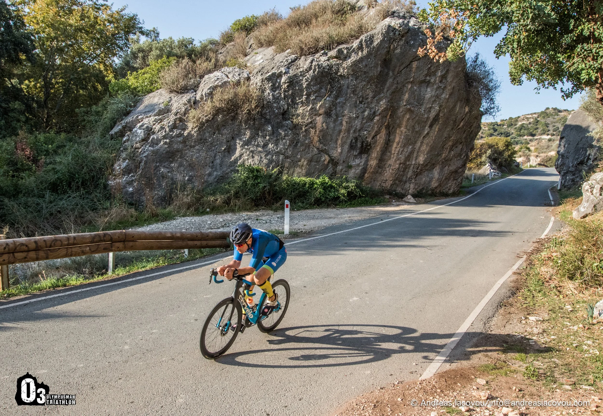A cyclist in a blue and yellow outfit riding a black bicycle on a winding mountain road surrounded by large rocks and greenery under a clear sky.