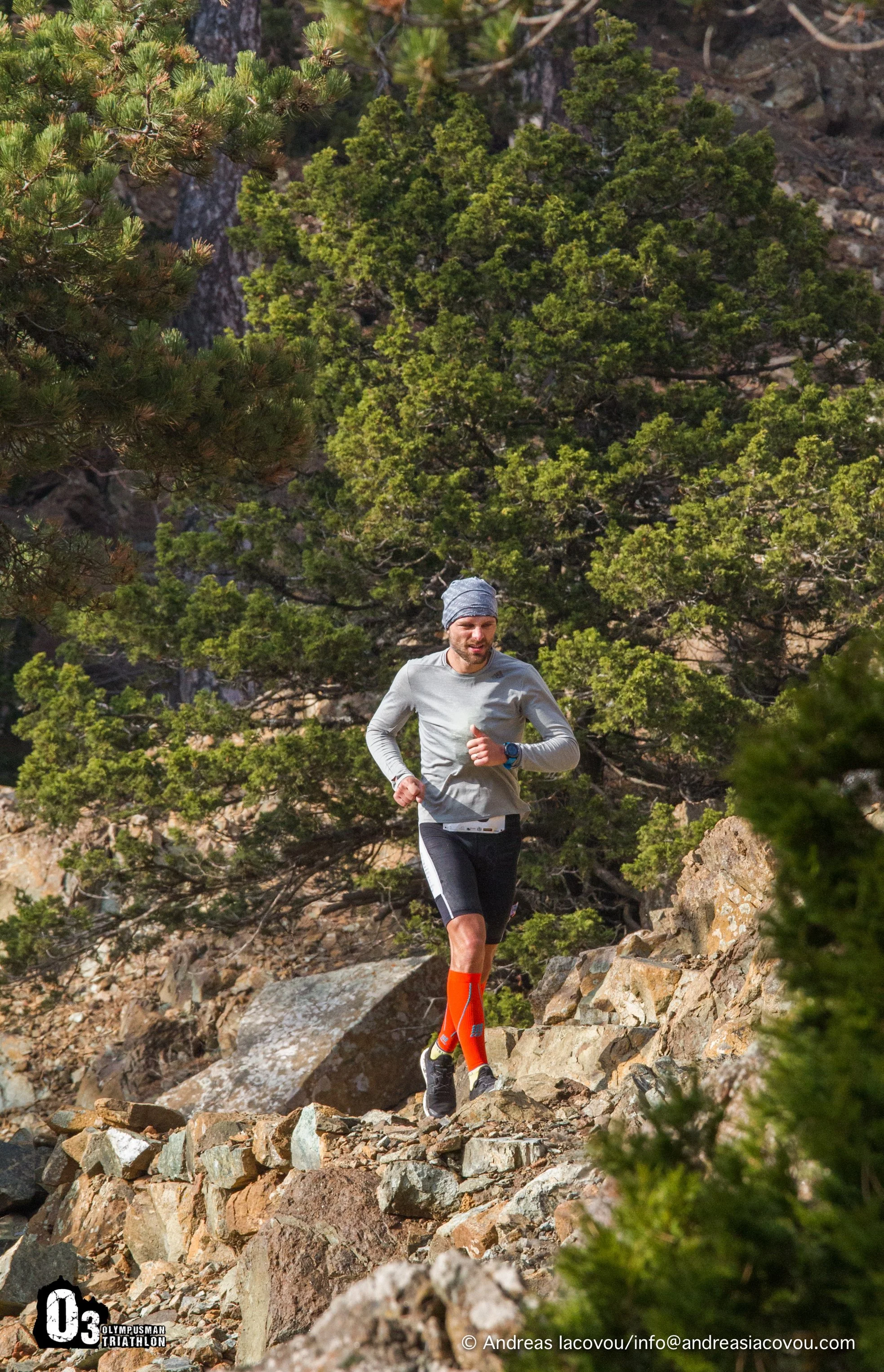 A man dressed in athletic wear, including a gray long-sleeve shirt, black shorts, orange socks, and a gray beanie, is running on a rocky trail through a wooded area with green trees and large rocks.