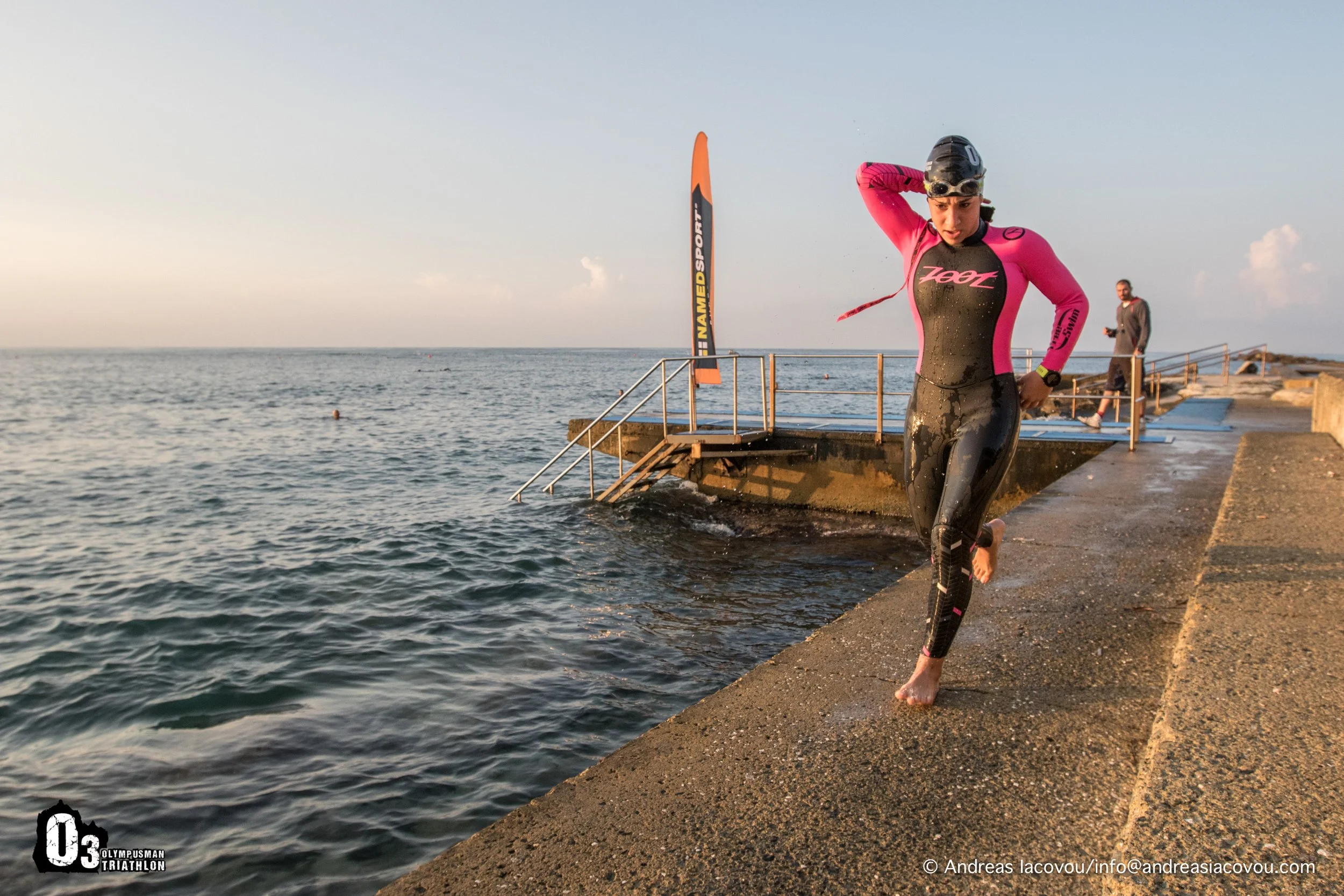 Female triathlon athlete in a pink and black wetsuit, wearing a swim cap and goggles, exiting the water onto a concrete dock at the ocean during sunrise or sunset.