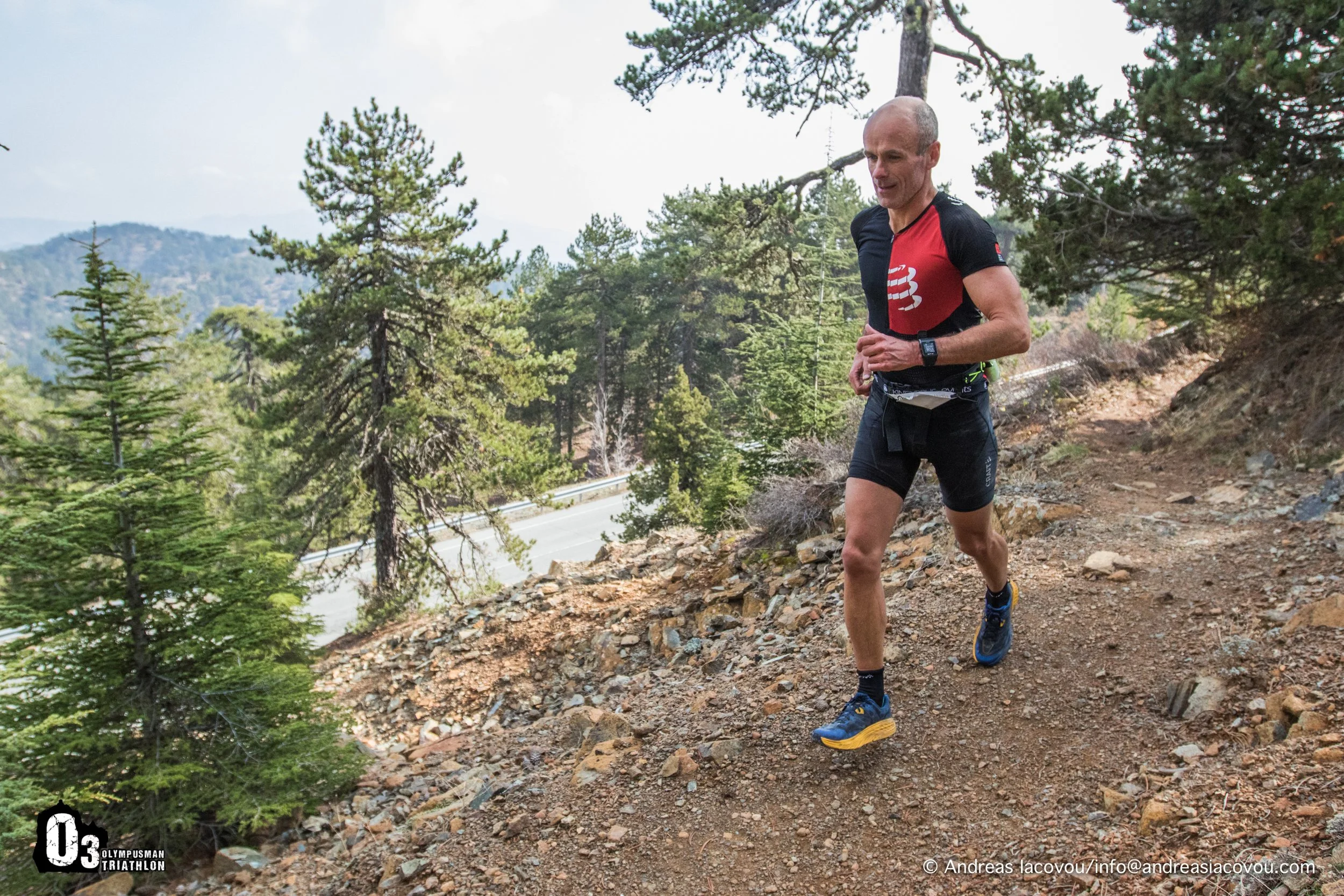 A man running on a rocky mountain trail surrounded by pine trees during daytime.