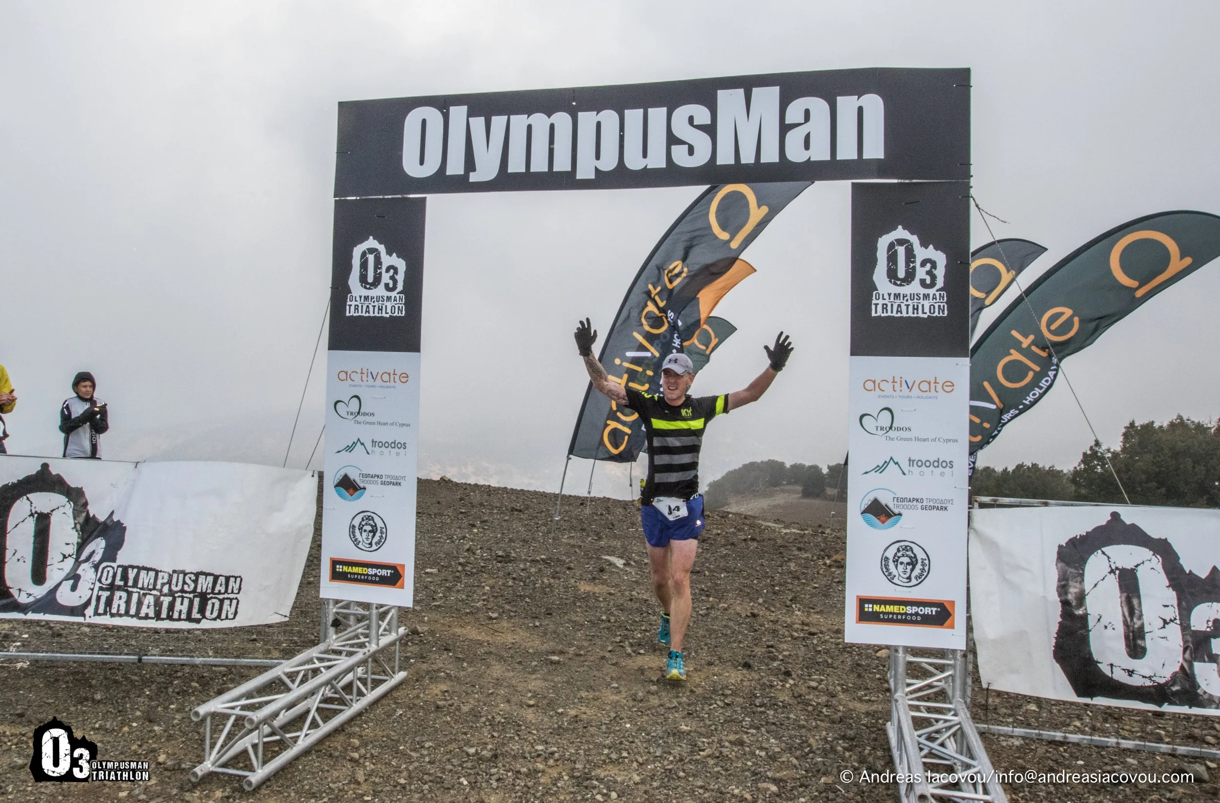 Triathlete crossing the finish line at OlympusMan triathlon event, smiling with arms raised, on a rocky outdoor terrain with banners and flags.