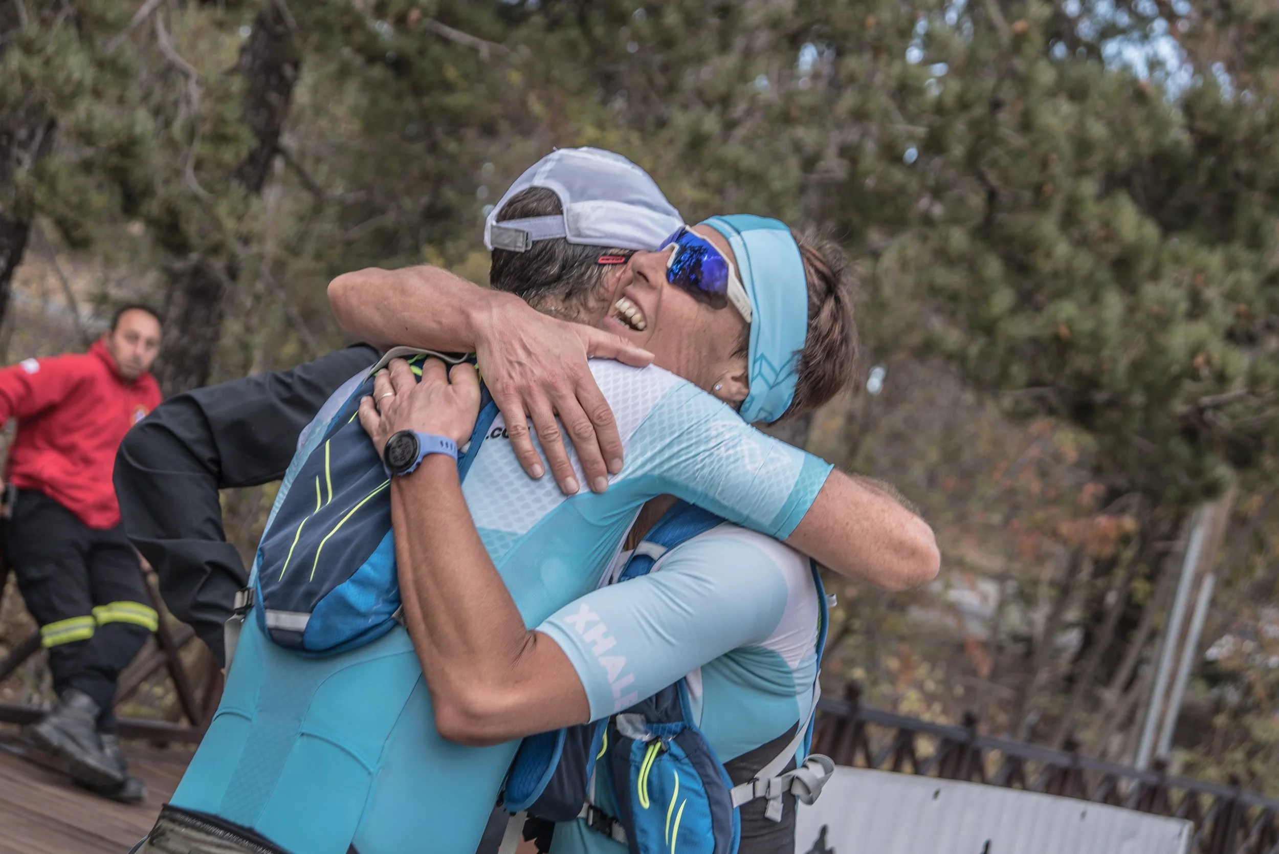 Two people hugging each other with smiles, outdoors in a forested area, after a sporting event or race. One person wears a light blue shirt, cap, and sunglasses, and the other wears a long-sleeve black shirt.