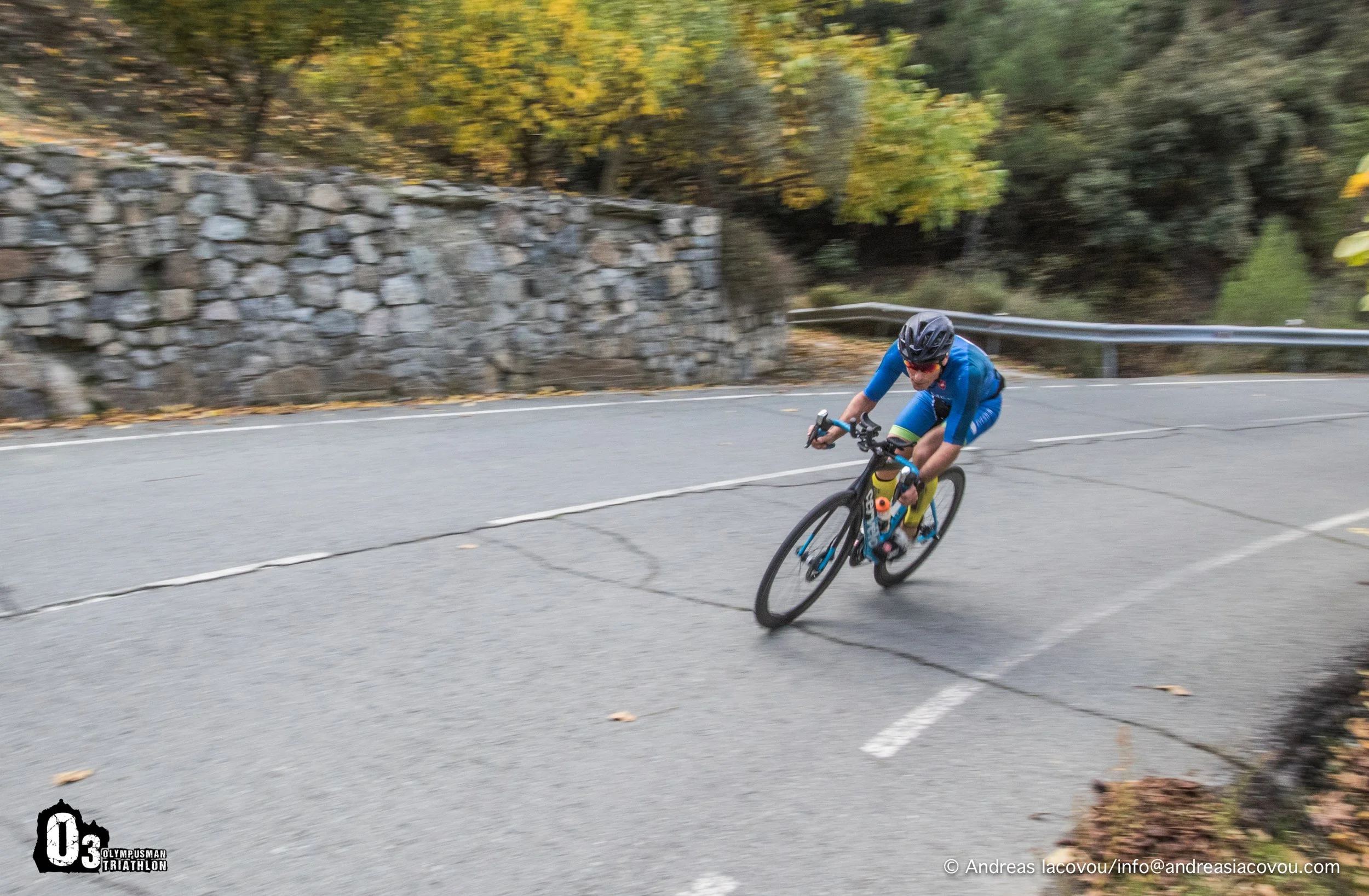 A cyclist in a blue outfit and helmet leaning into a turn on a mountain road with guardrails and trees in the background.