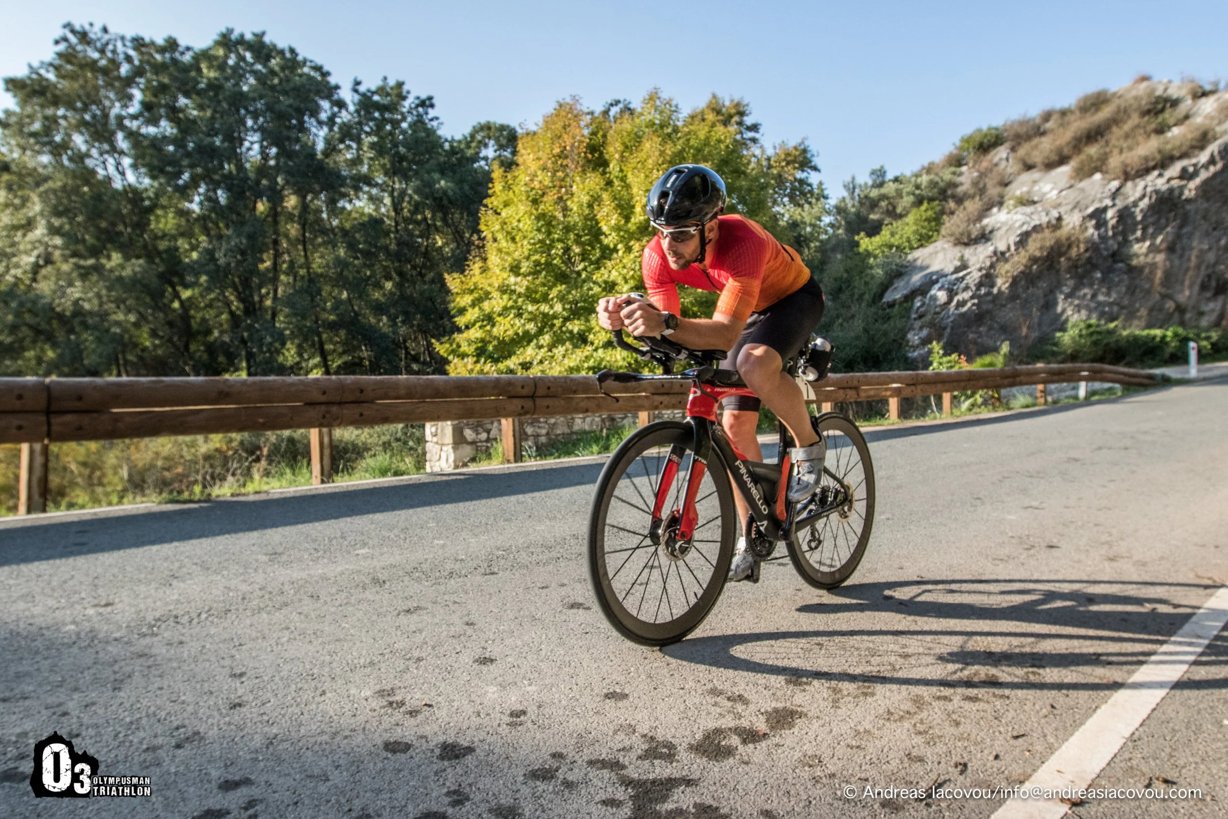 A male cyclist wearing a black helmet, sunglasses, a red and orange shirt, and black shorts rides a black and red road bike on a paved mountain road with a wooden guardrail. Trees and rocky hills are in the background.
