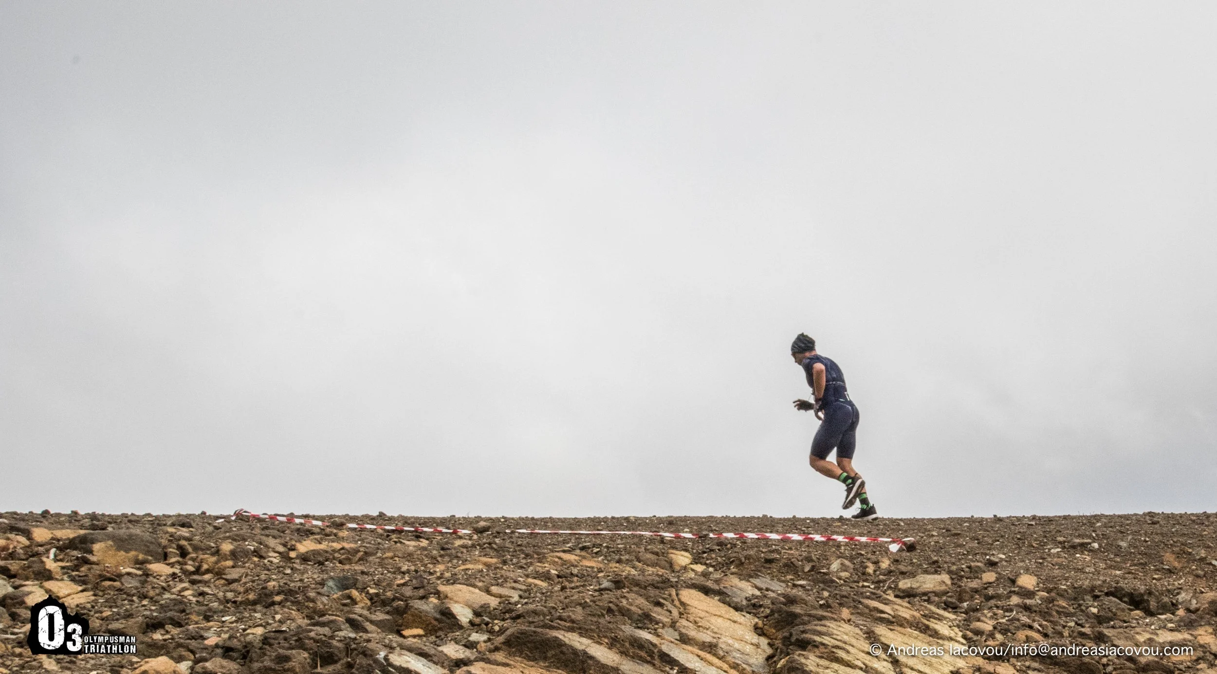A female athlete running on a rocky, barren terrain under a cloudy sky during a triathlon event.