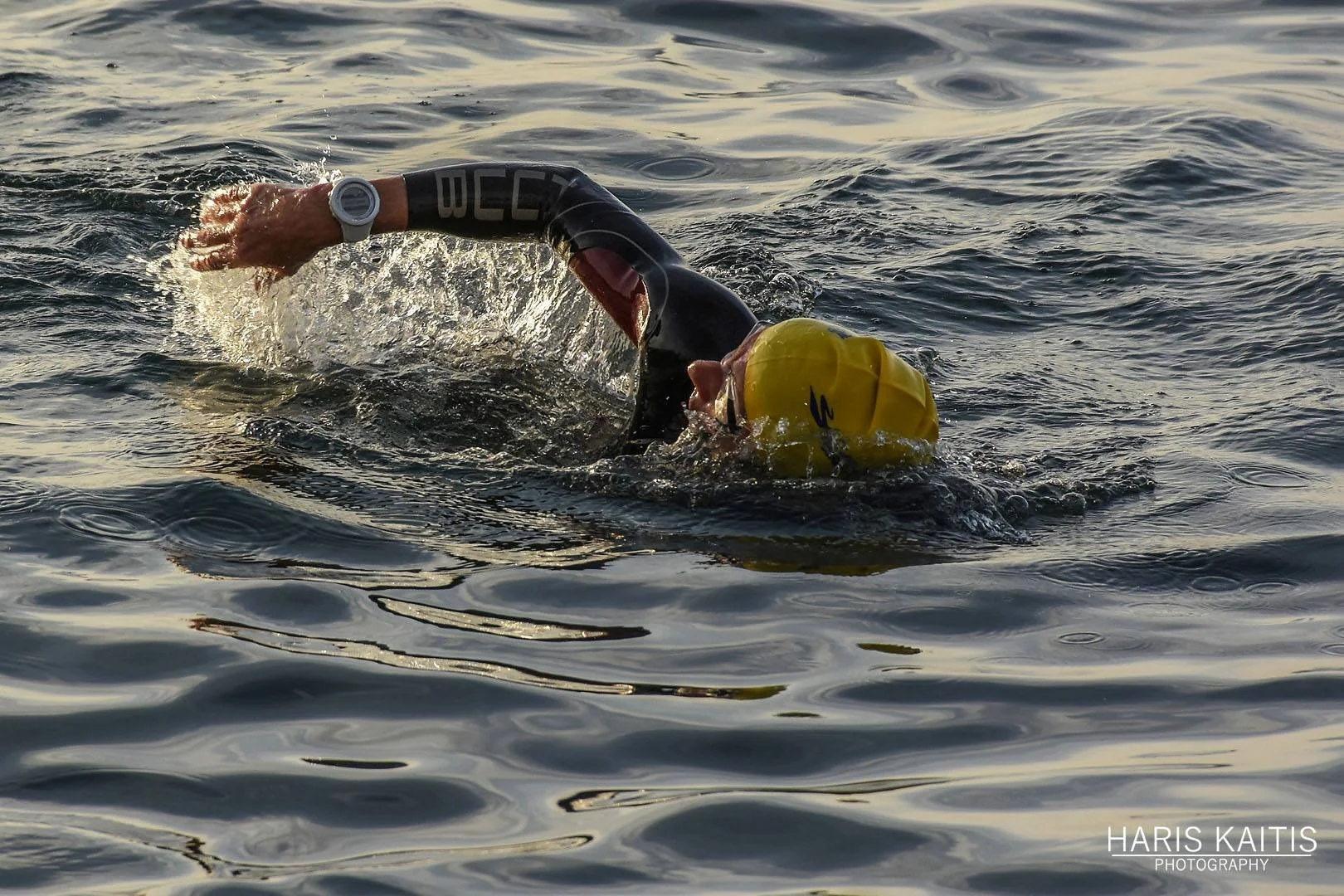A person swimming in open water, wearing a yellow swim cap, a black wetsuit with red accents, and a white wristwatch, captured during daytime with rippling water around them.