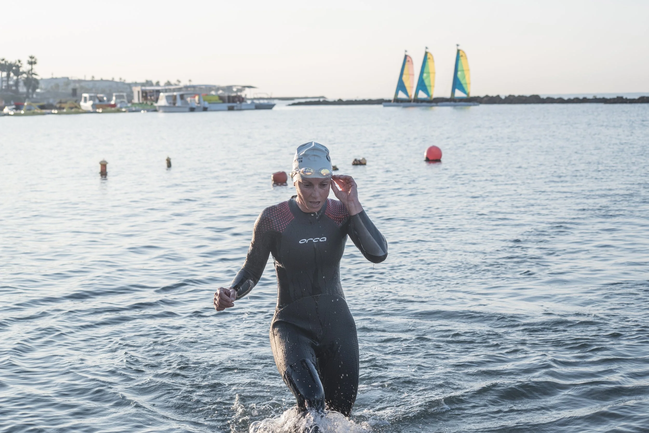 A woman in a black wetsuit and swim cap exiting the water during a triathlon or open water swim race, with sailboats, boats, and buoys visible in the background along a body of water.