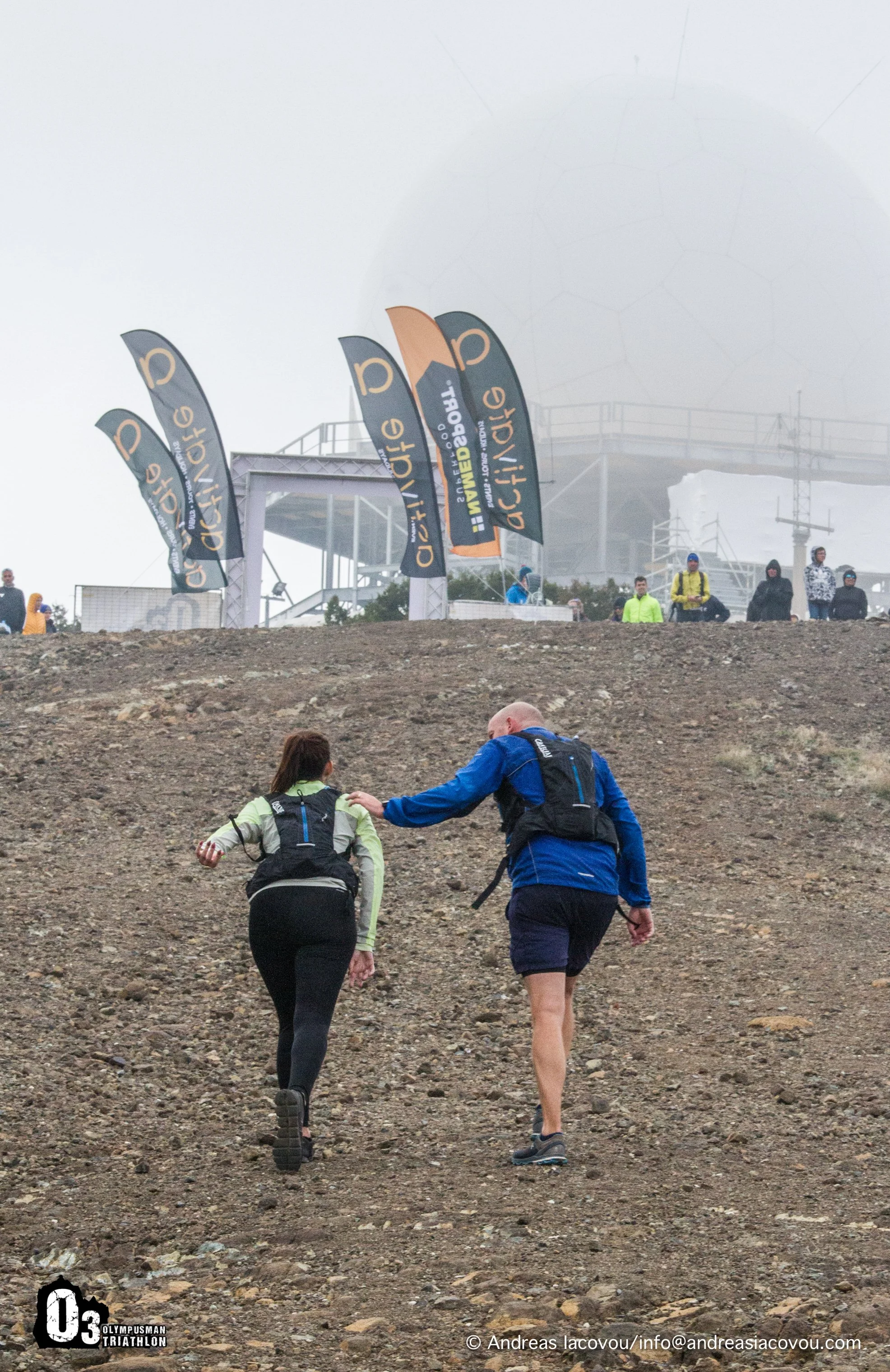 Two athletes with backpacks walking uphill on a rocky trail during a mountain race, with flags and spectators in the background in foggy weather.