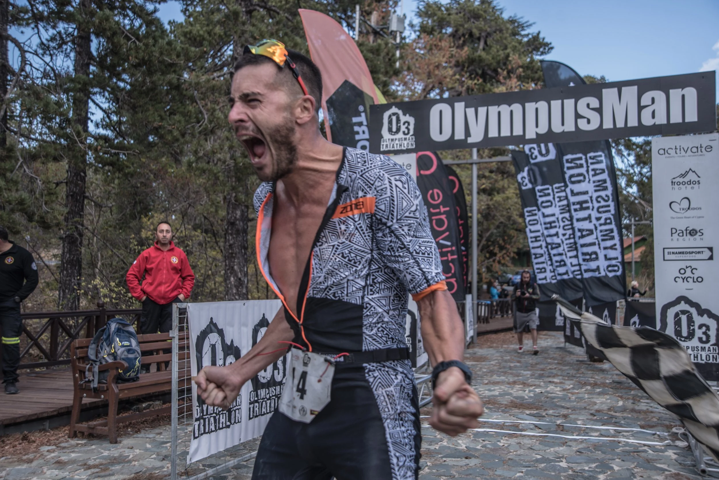 Male athlete crossing finish line at OlympuspMan triathlon, wearing bib number 4, with a fierce expression, clenched fists, and an open mouth in a victory pose, surrounded by race banners and spectators.