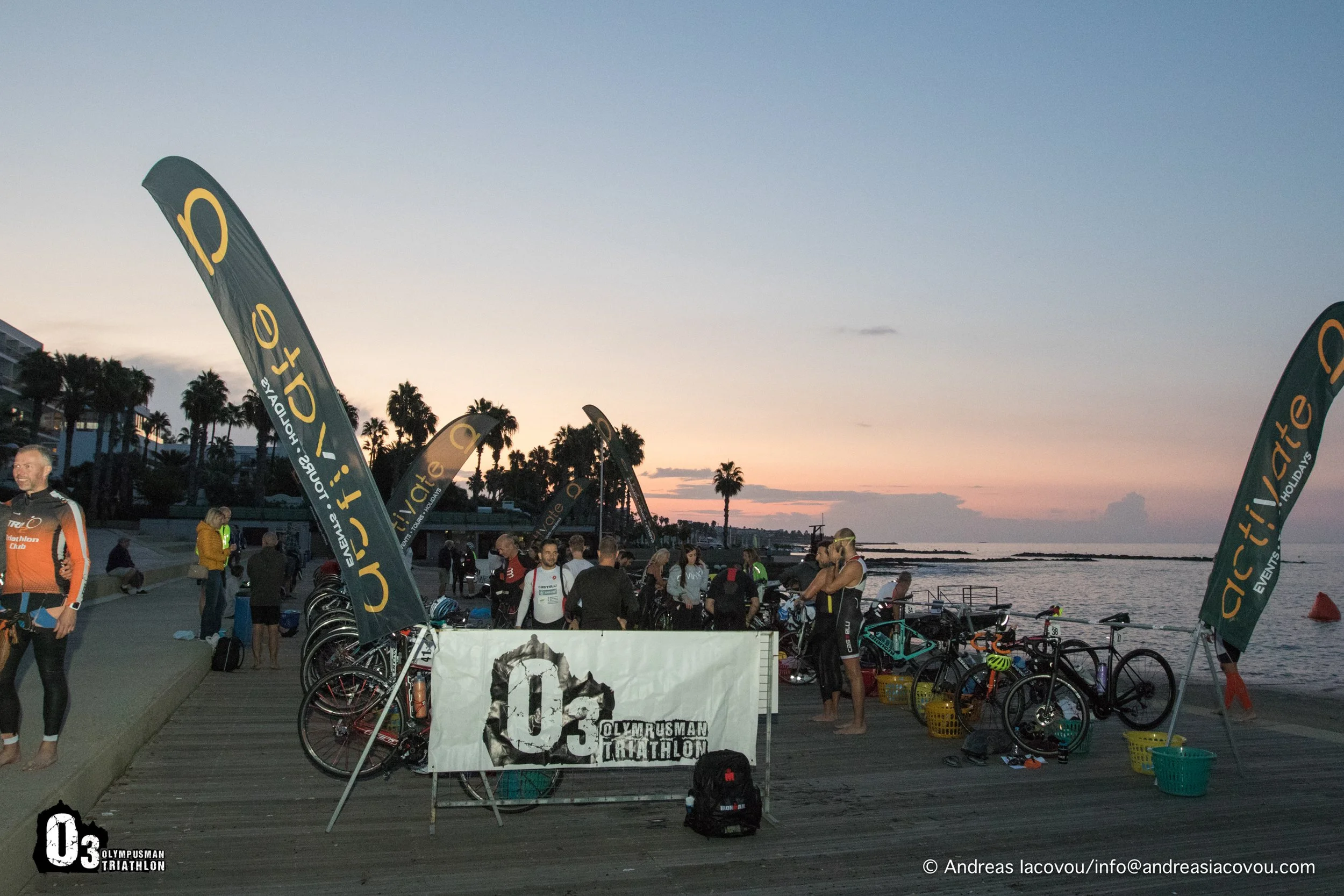 Triathlon event by the water at sunset with bikes, participants, and flags reading 'Active Fit'.