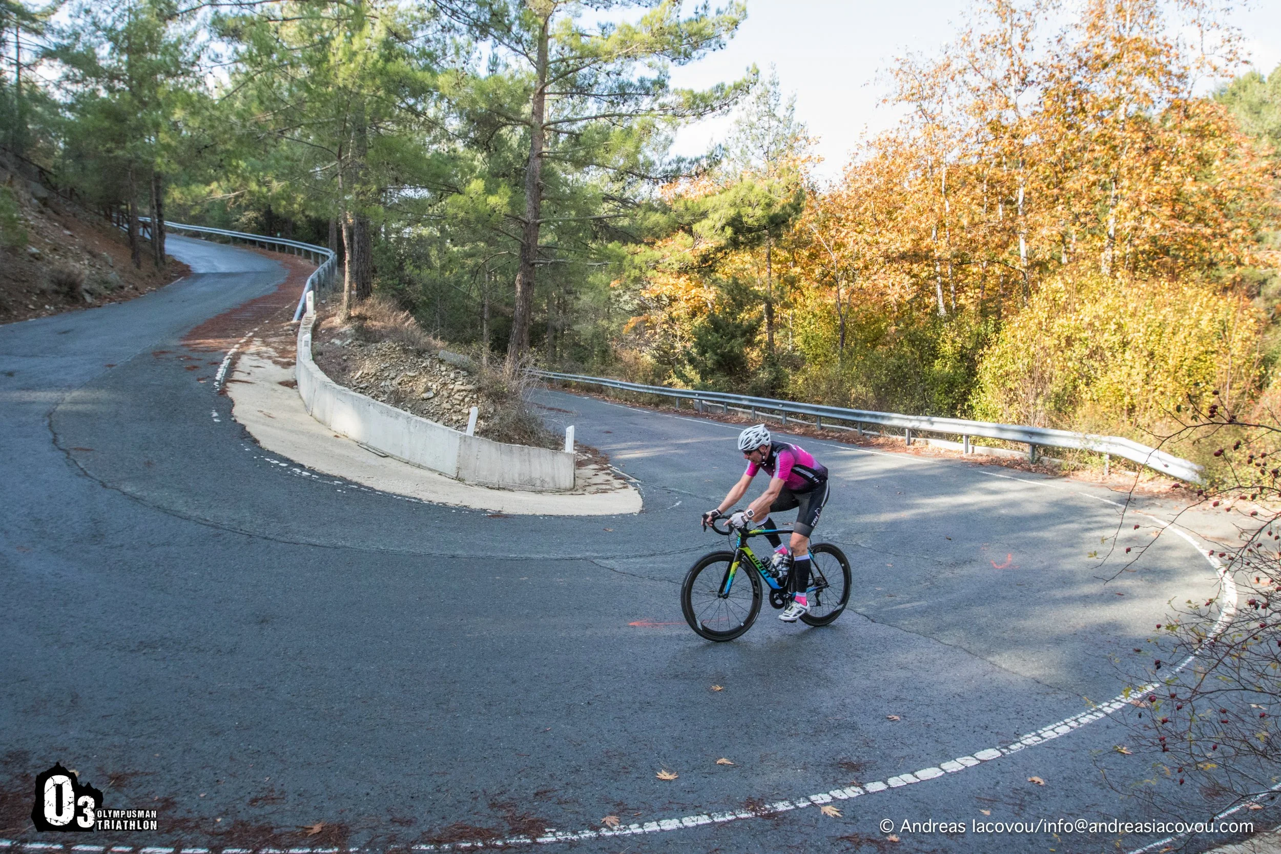 A cyclist wearing a pink jersey and a white helmet riding on a winding mountain road surrounded by trees with fall foliage.