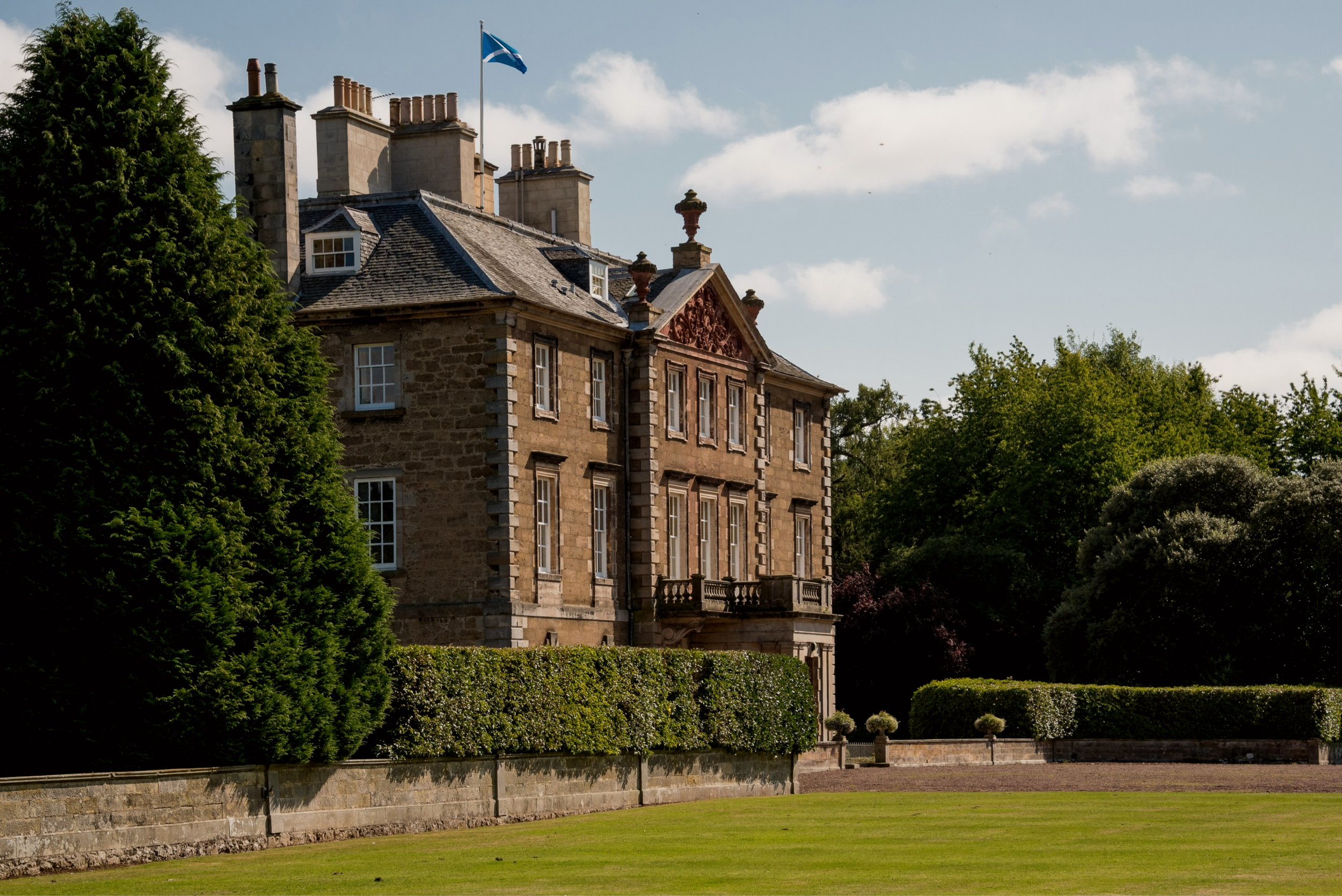A large historic stone house with multiple chimneys, windows, and a balcony, surrounded by green trees and a manicured lawn, with a Scottish flag flying on top.