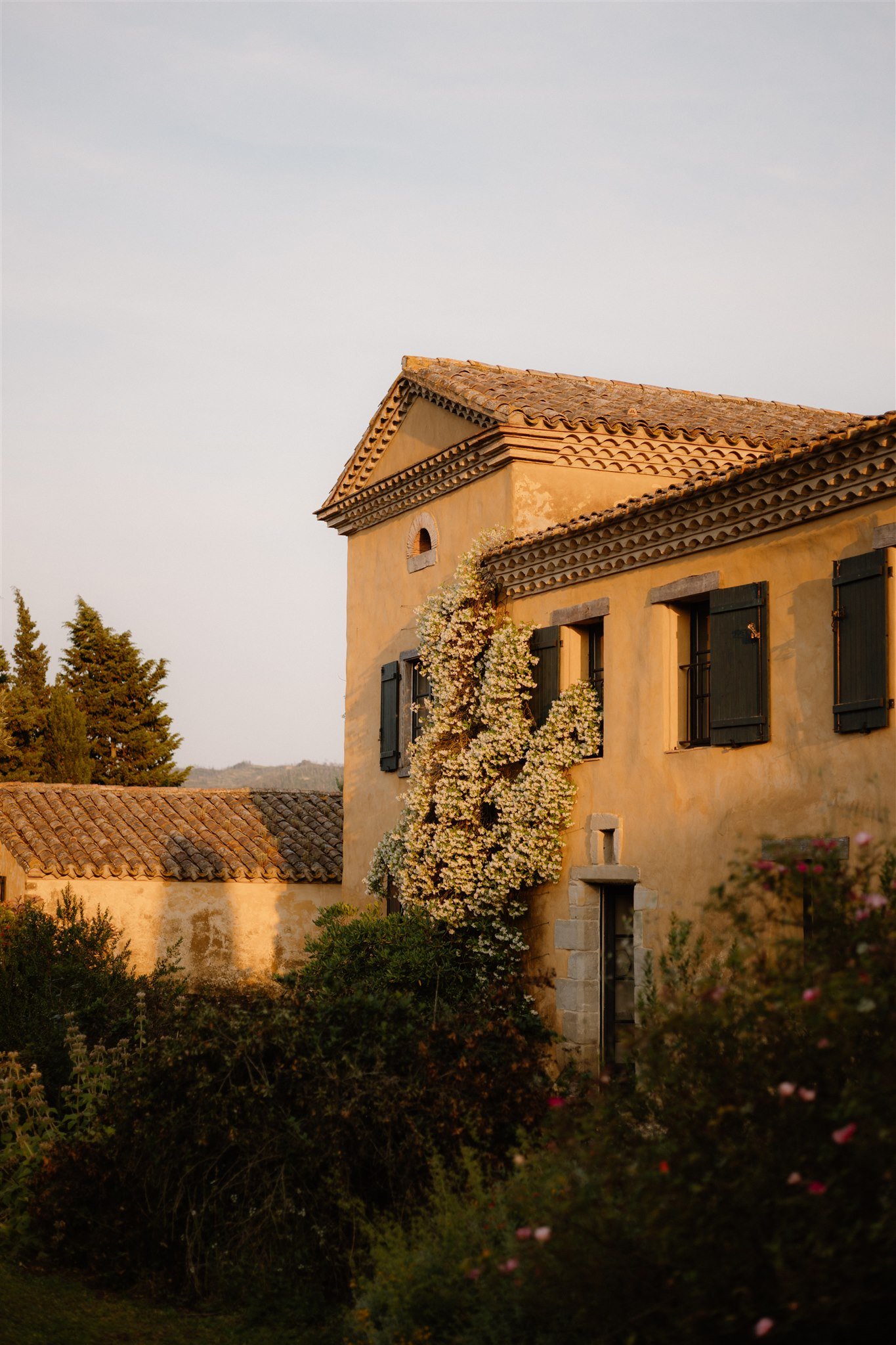 An old Mediterranean-style house with yellow stucco walls, green shutters, and a tiled roof, surrounded by lush greenery and flowering plants during sunset.
