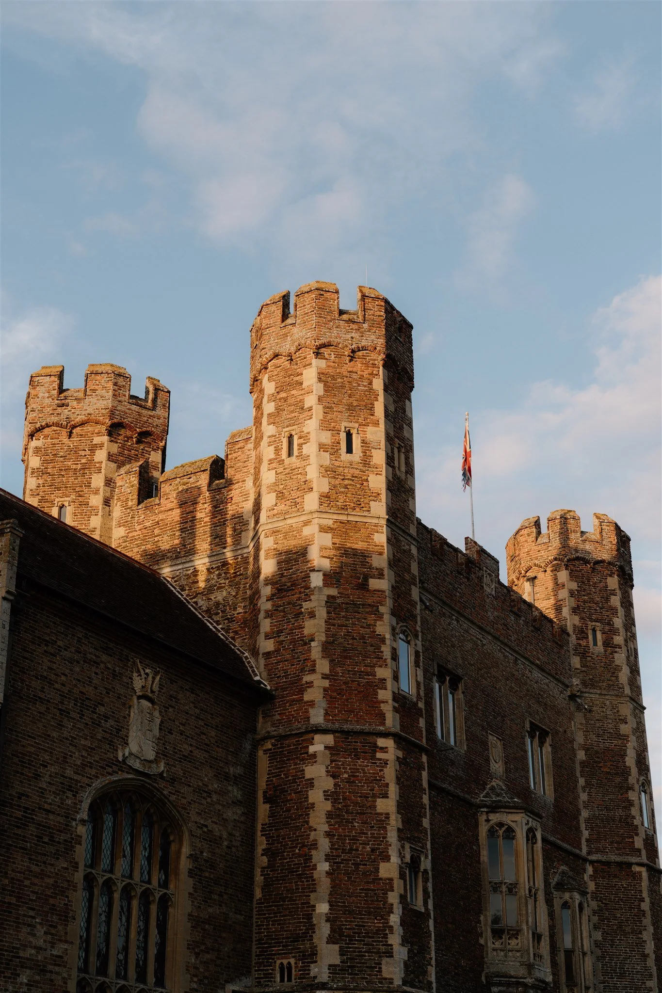 A historic brick castle with multiple towers, crenellations, and a Union Jack flag flying from a flagpole, set against a blue sky with scattered clouds.