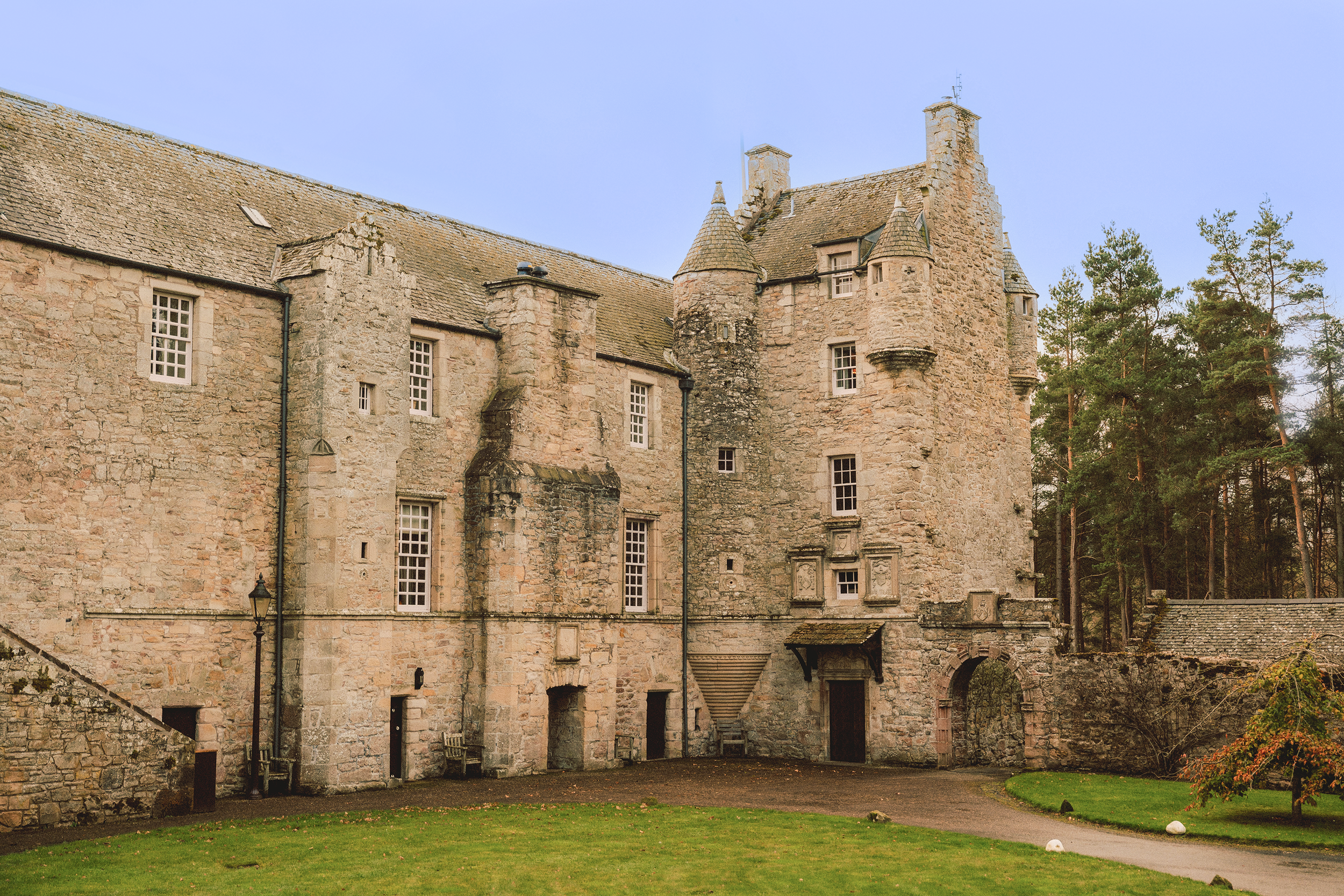 A historic stone castle with multiple towers and small windows, surrounded by a grassy lawn and pine trees, under a clear blue sky.