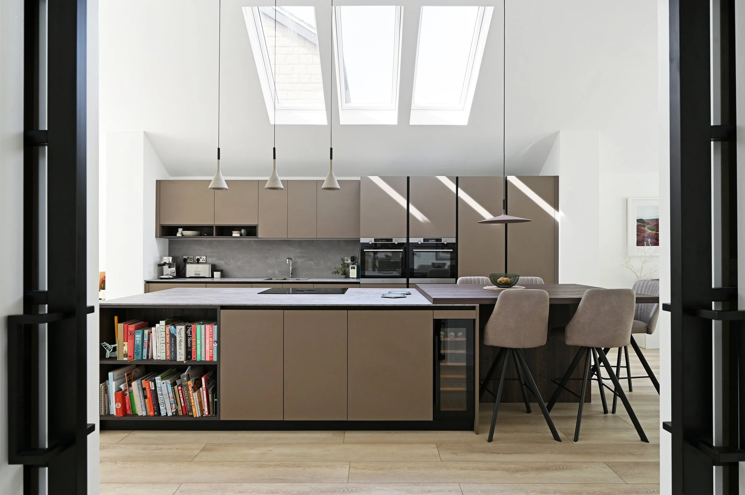 Modern kitchen with beige cabinets, a large island with a gray countertop, and a breakfast bar with four beige chairs. Above, three pendant lights hang, and there are skylights on the ceiling allowing natural light. The background features built-in o