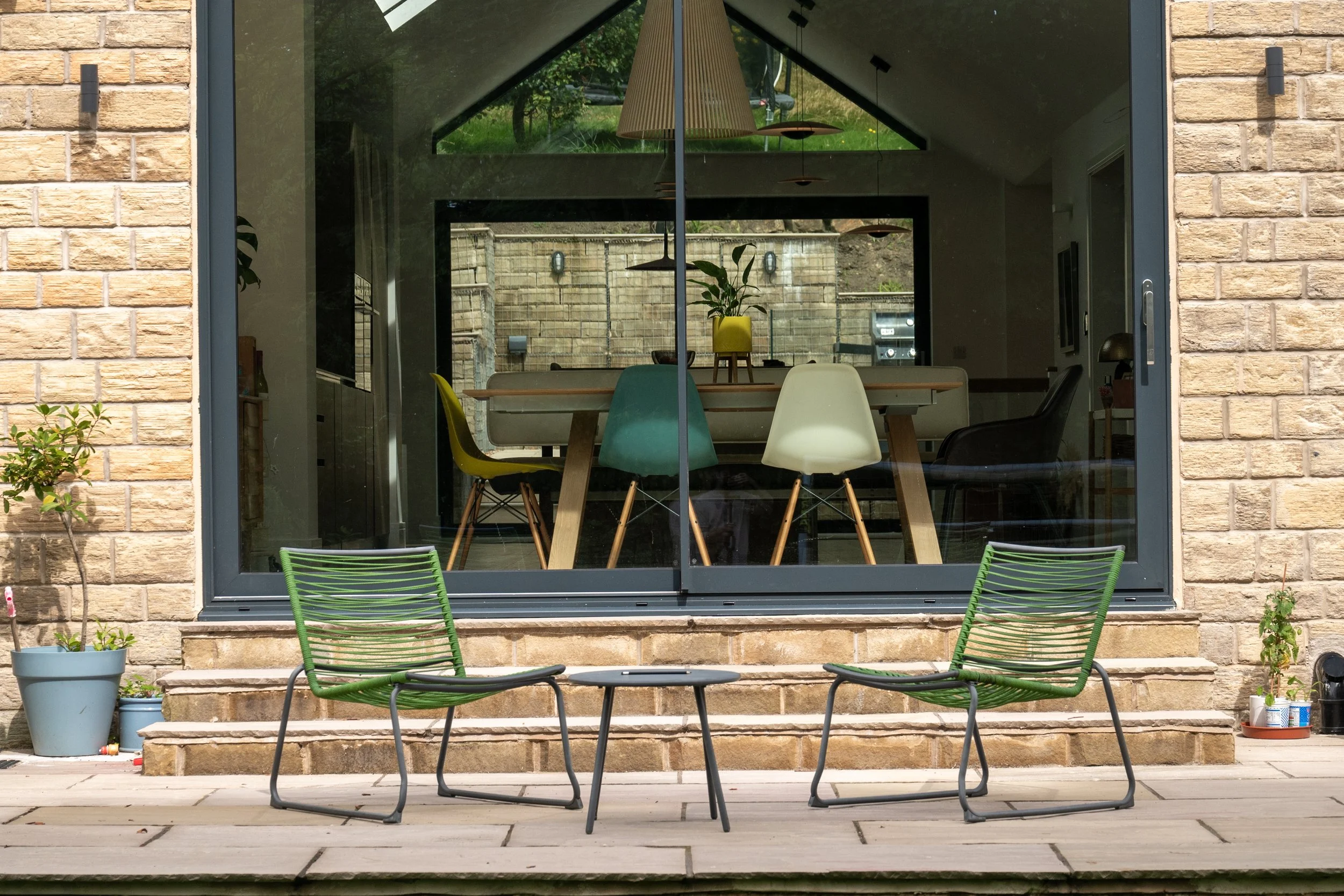 View of a house's patio with two green outdoor chairs and a small green table in front of a large sliding glass door, showing a dining room with colorful chairs and a potted plant inside.