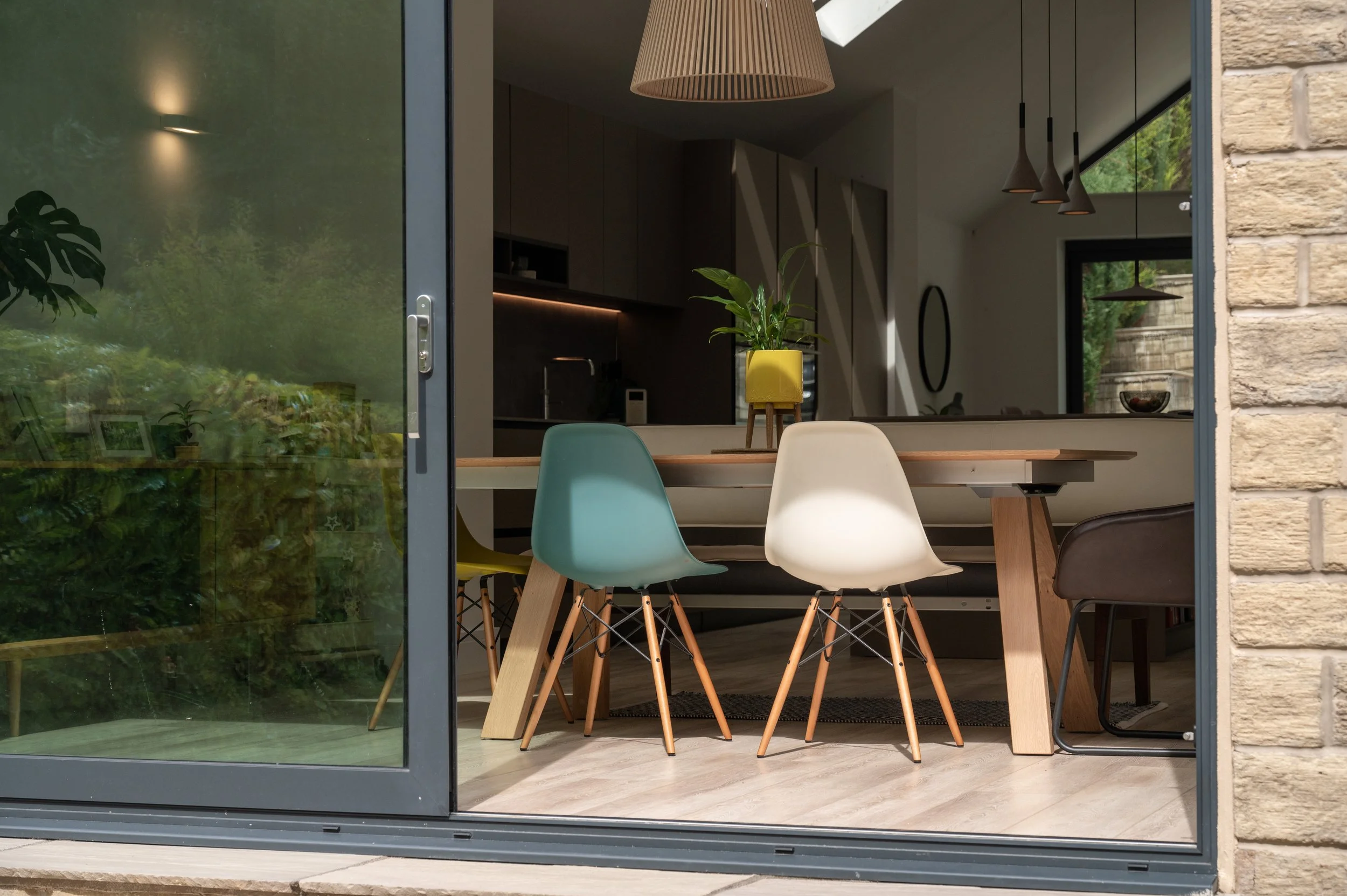 Modern dining room with a wooden table, colorful chairs, and a potted plant on the table, viewed through a sliding glass door.