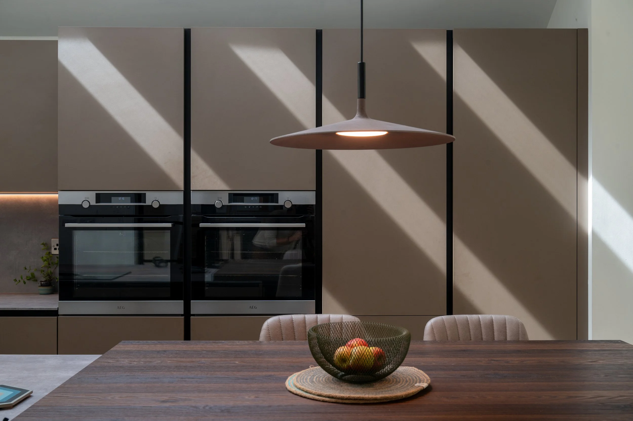 Modern kitchen with built-in ovens, beige cabinetry, a wooden dining table with a bowl of apples, and a hanging pendant light.