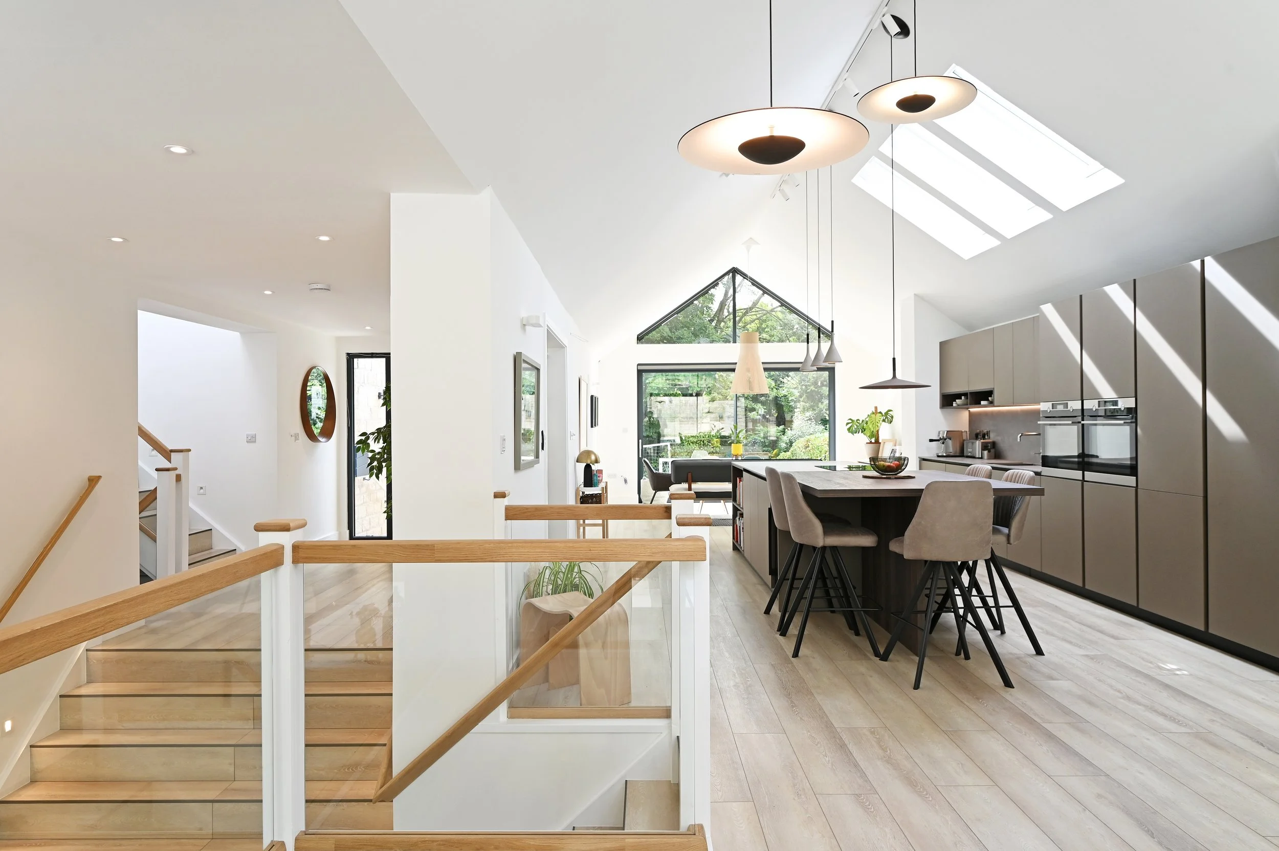 Open-concept kitchen and living room with modern design, neutral tones, large windows, skylights, a kitchen island with chairs, and wooden flooring.