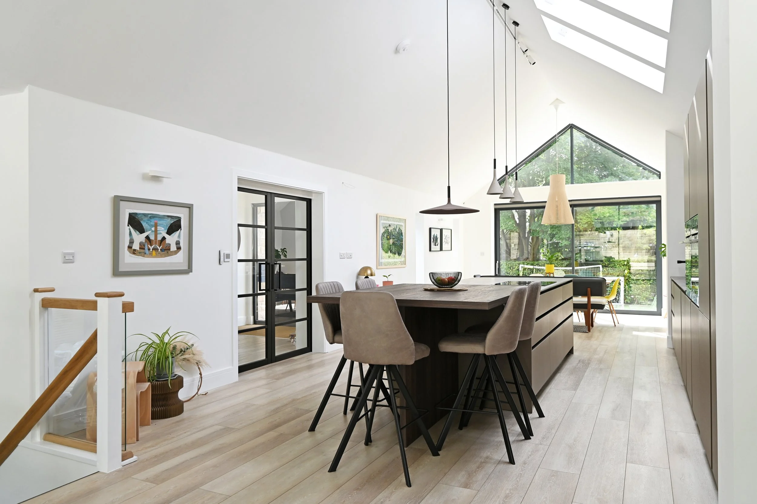 Modern open-concept kitchen and dining area with white walls, light wooden flooring, a central island with seating, pendant lights, large windows, and artwork on the walls.