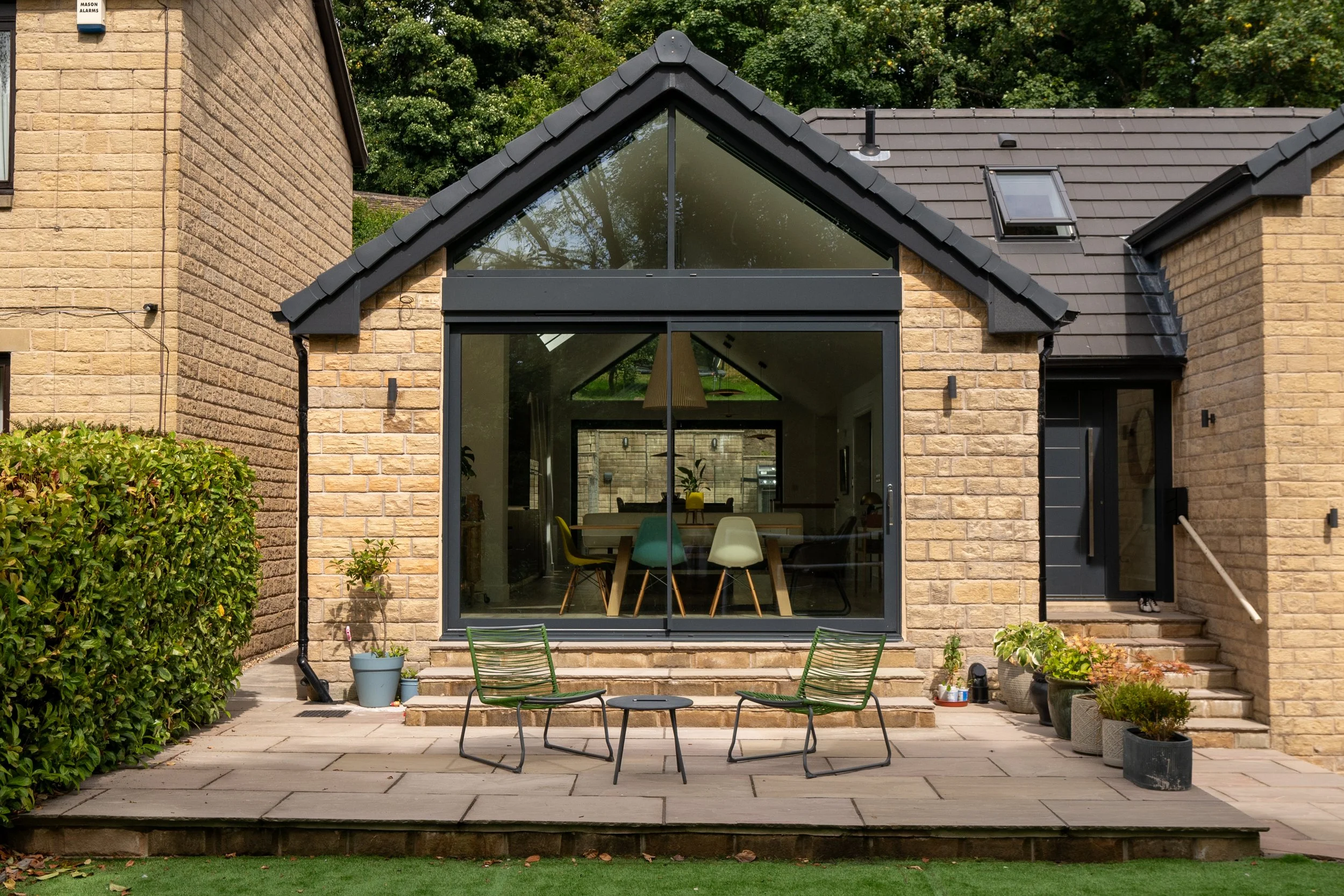 View of a modern brick house with a large glass window and sliding door leading to an outdoor patio with two metal chairs and a small table, potted plants along the steps, and trees in the background.
