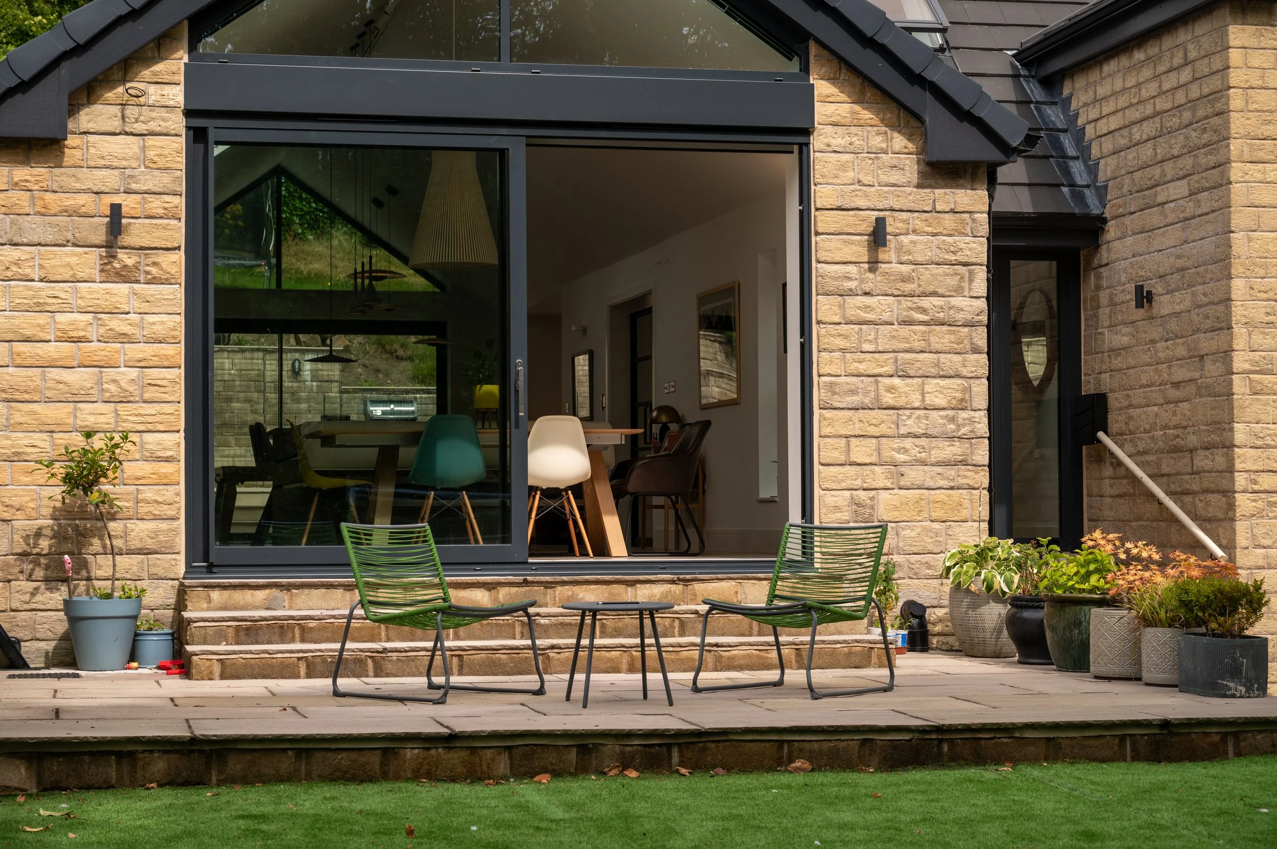 View of a backyard patio with two green chairs and a small table, outdoor potted plants, and a brick house with a large sliding glass door leading inside to a dining area.