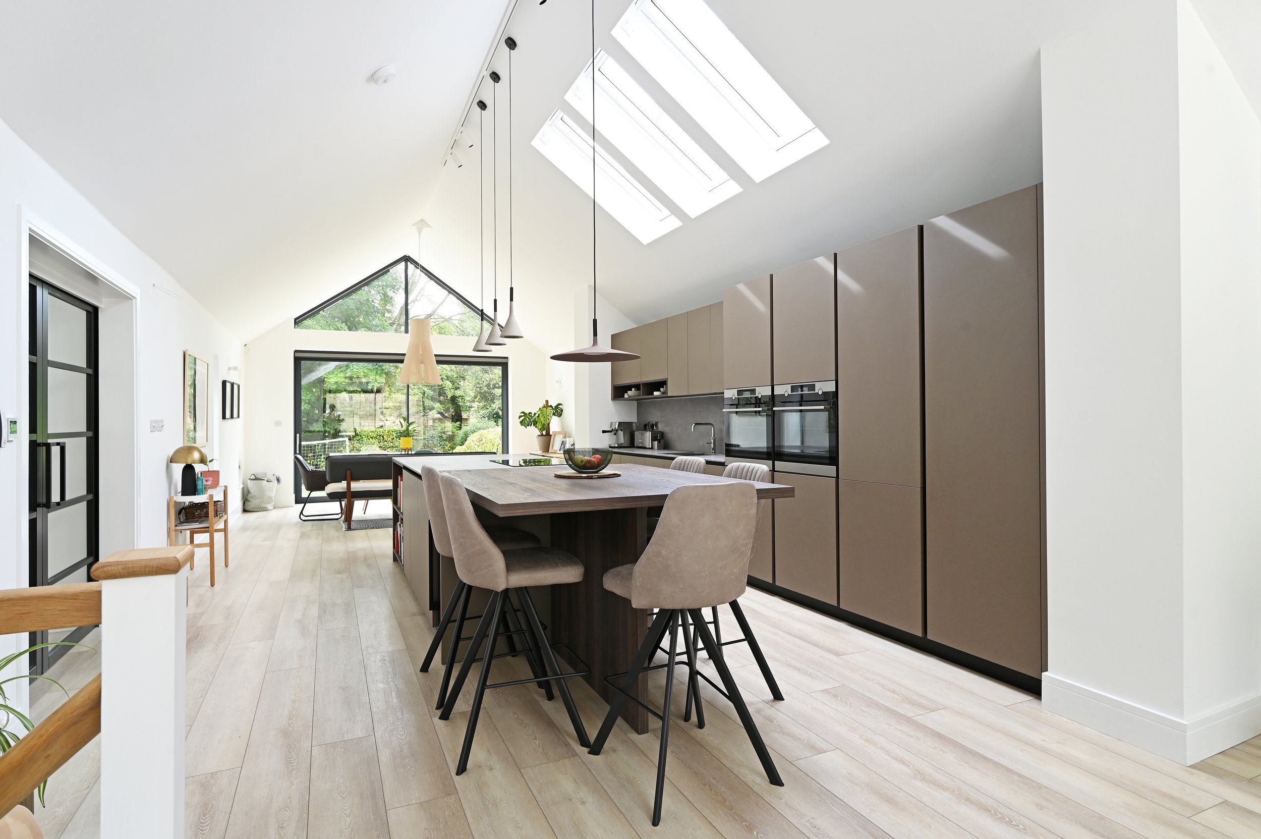 Modern open-concept kitchen and dining area with natural light from skylights and large windows, beige cabinetry, a central kitchen island with seating, and a sitting area with a large window and garden view.
