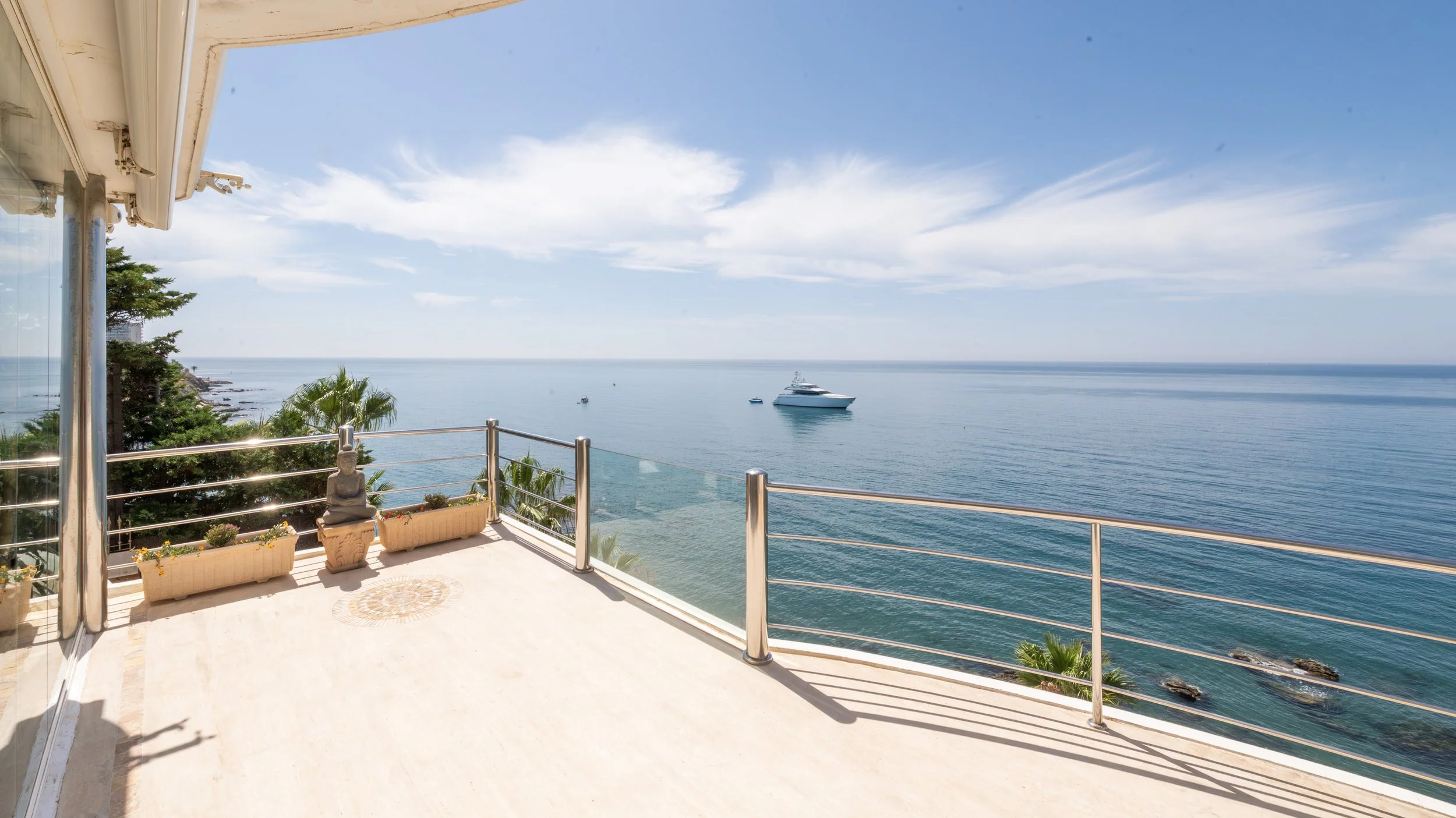 Vista panorámica del mar desde un balcón con barandilla de metal, con un yate grande y algunos barcos más pequeños en el agua, cielo despejado con nubes ligeras, plantas y decoración en la terraza.