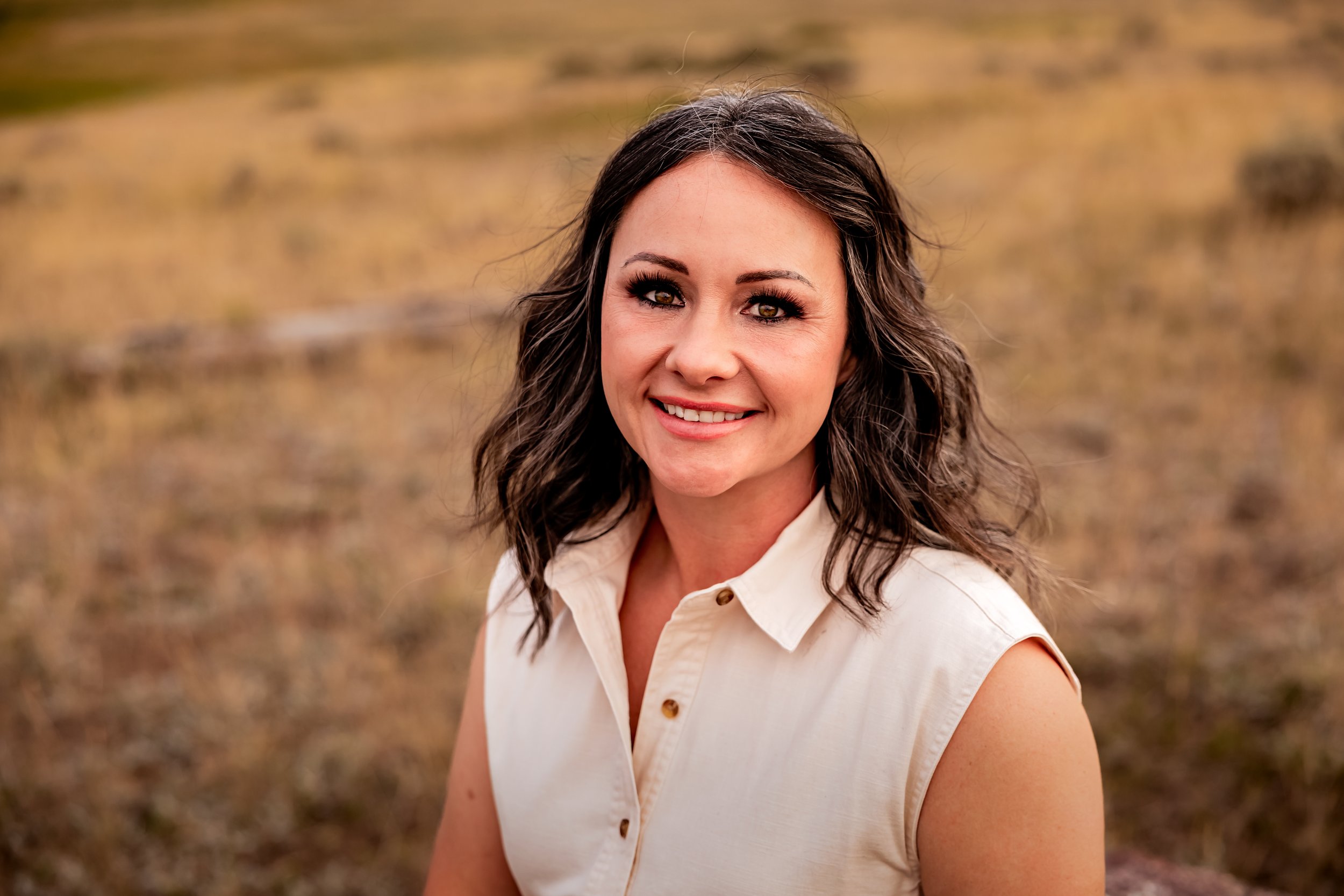 A woman with shoulder-length wavy dark hair and light makeup smiling outdoors with a blurry brown grass background.