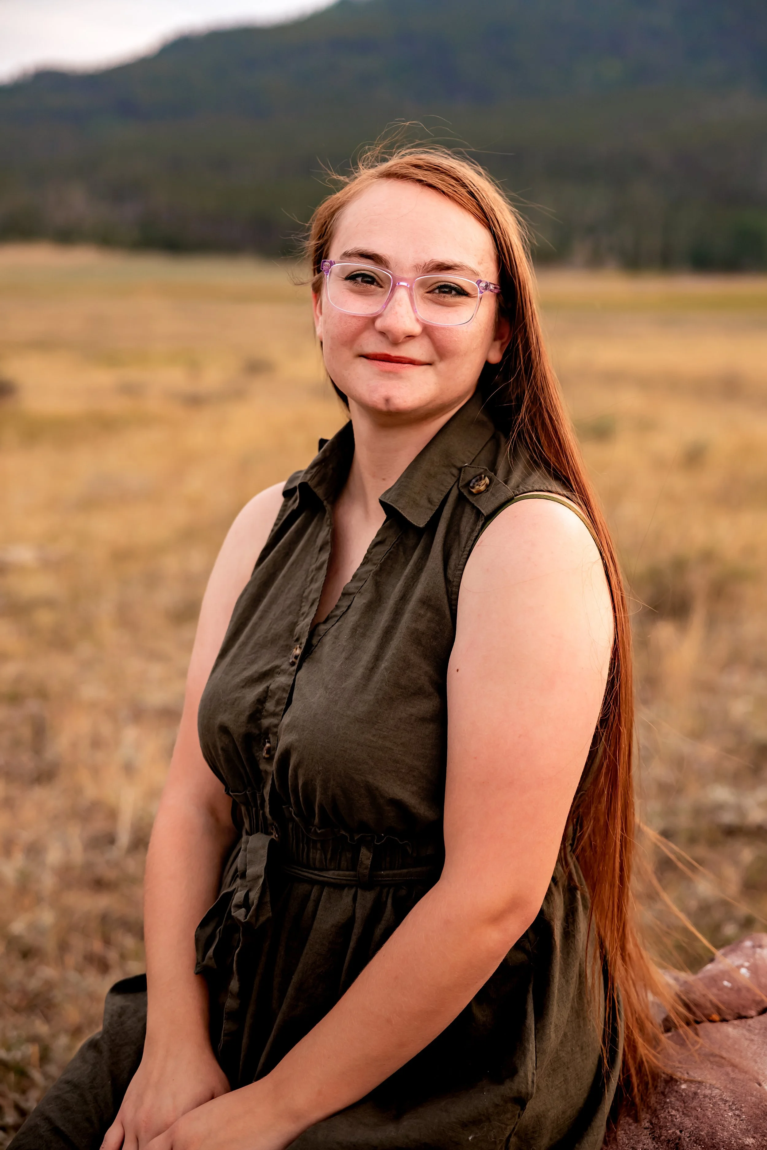 A woman with long red hair wearing glasses and a sleeveless dark green dress sits outdoors in a field with mountains in the background.