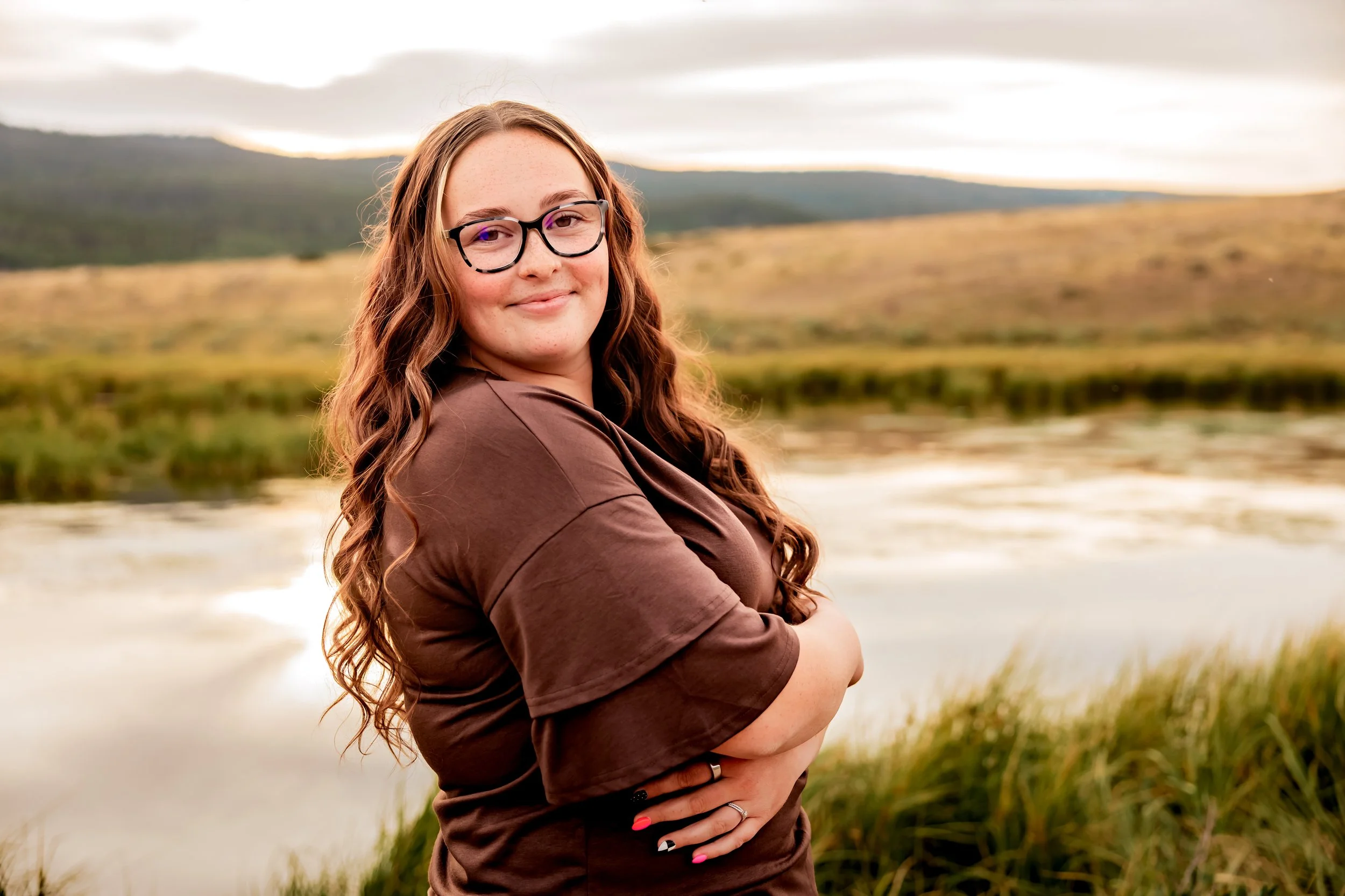 A woman with long curly hair and glasses standing outdoors near a body of water with hills in the background, smiling at the camera.