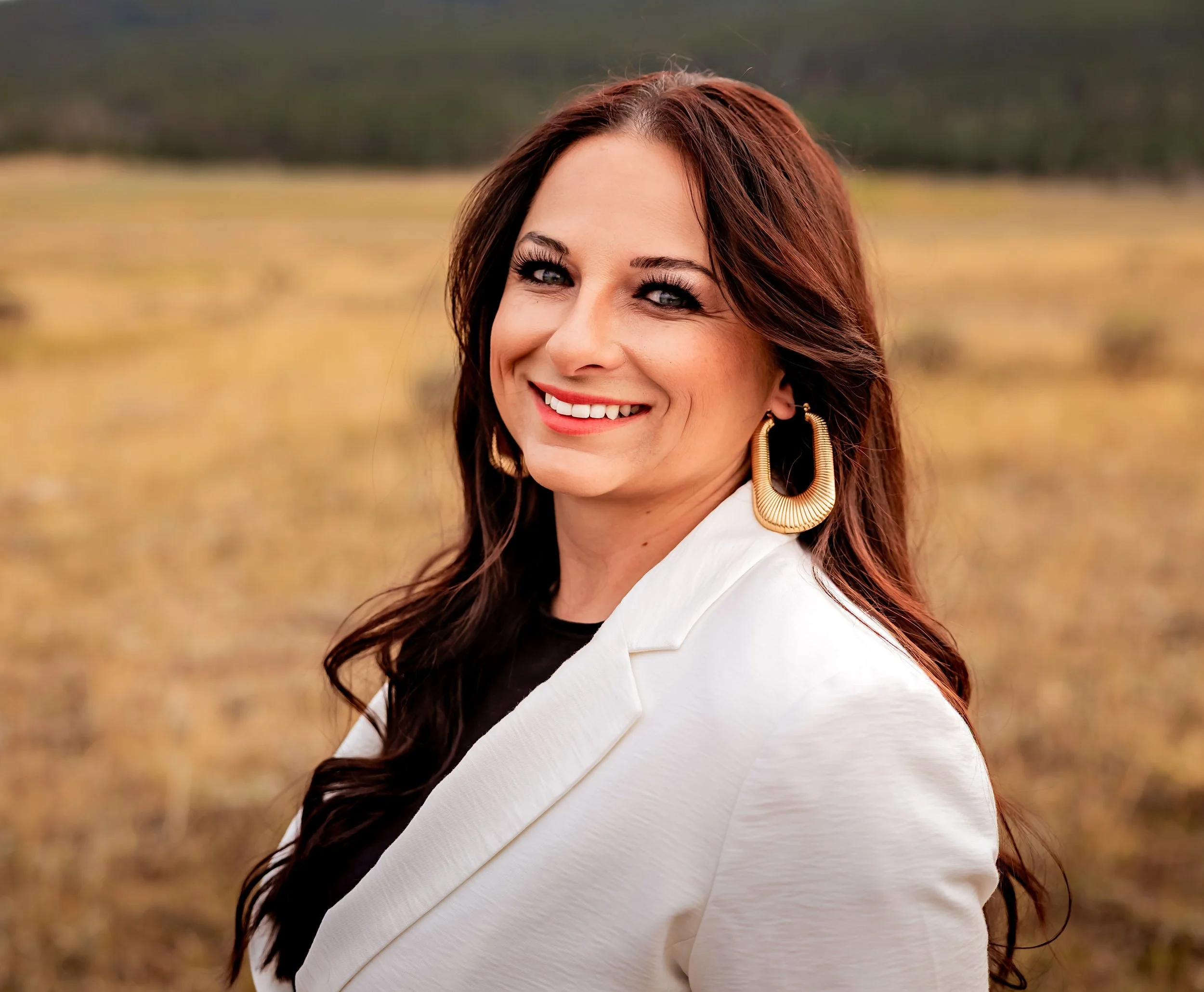 A woman with long brown hair and large gold earrings smiles outdoors in a field with a blurred background.
