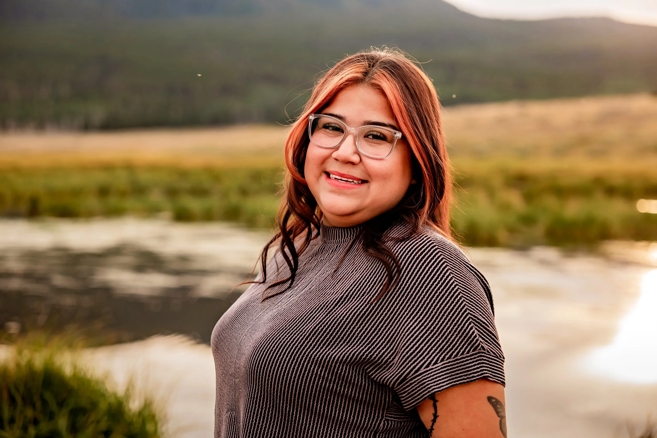 A woman with red hair, glasses, and a striped shirt smiling outdoors near a body of water with a blurred natural landscape in the background.