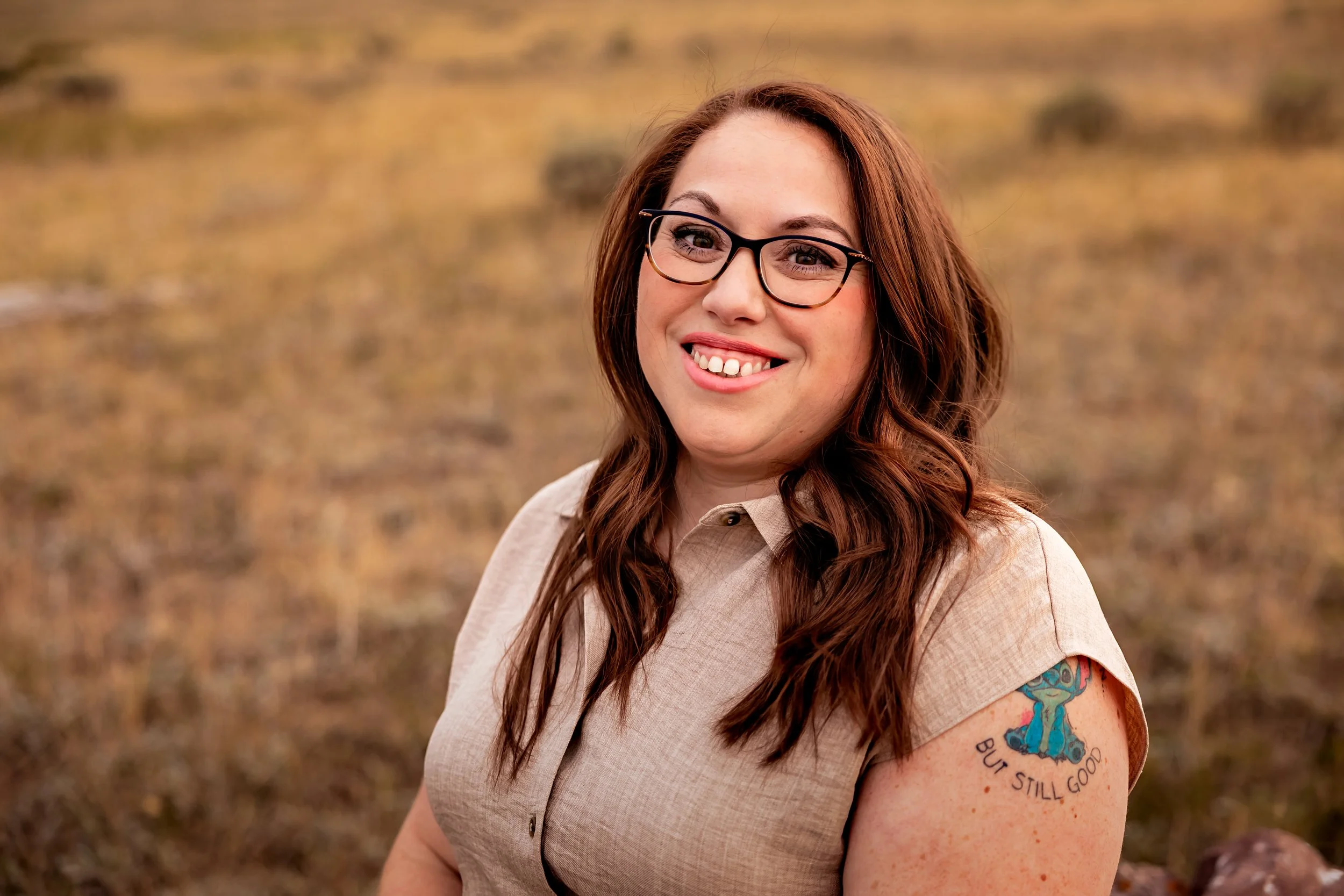 A woman with long reddish-brown hair, glasses, and a tan shirt standing outdoors in a grassy area, smiling with her tongue slightly out, showing a tattoo of a blue character on her upper arm with the words "But Still Good."