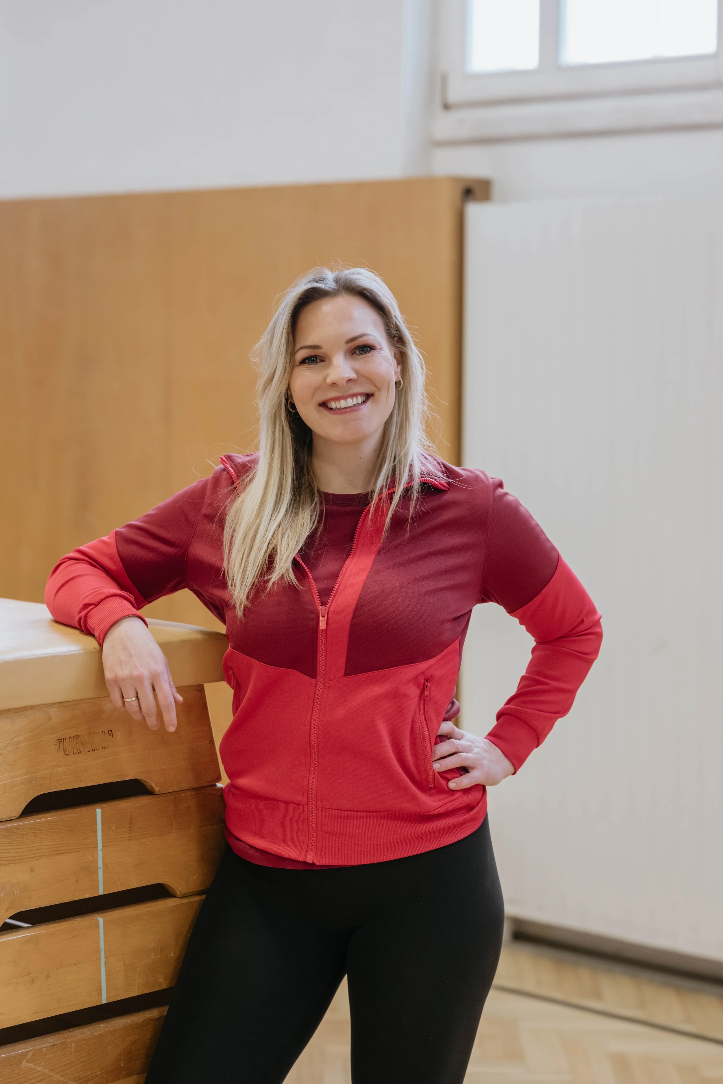 Frau mit blonden Haaren, rotem Sportjacke und schwarzen Hosen, lächelnd, in einem Raum mit Holzpolsterung und Fenster im Hintergrund.