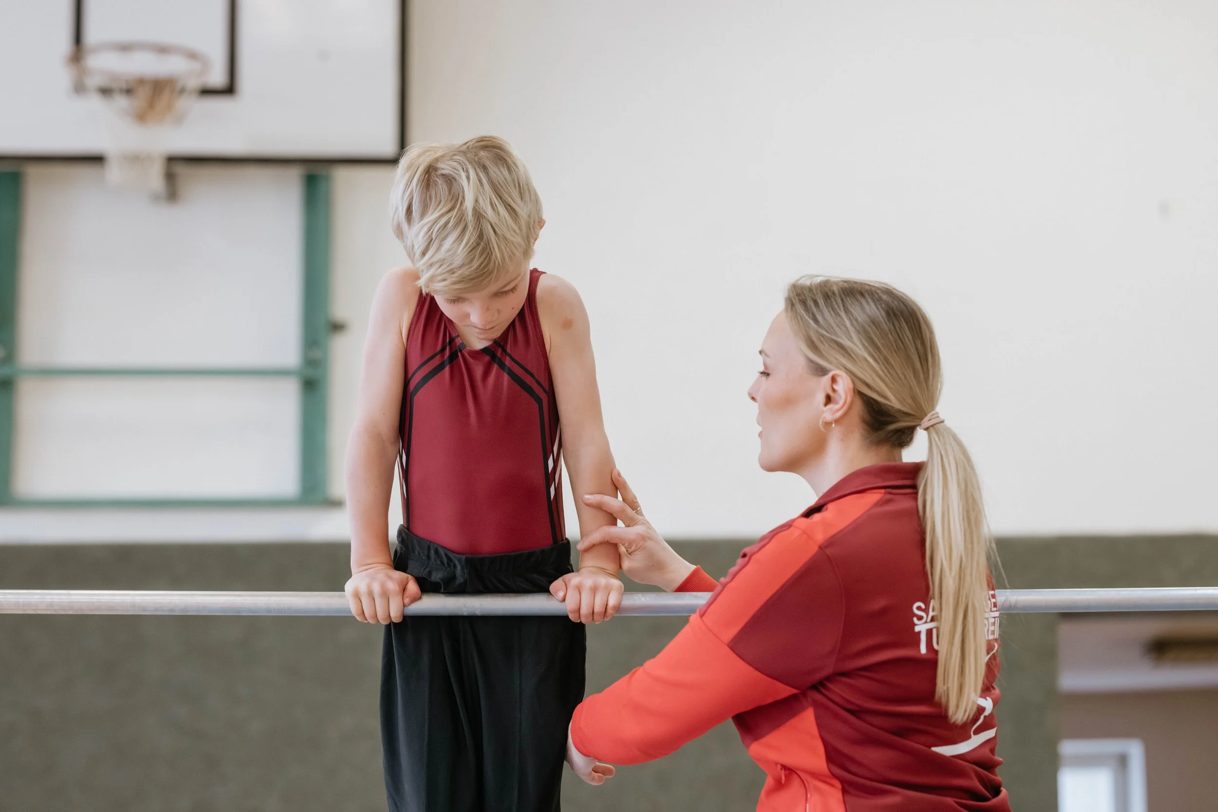 Ein Kind auf einem Barren im Turnunterricht, das von einer Betreuerin unterstützt wird.