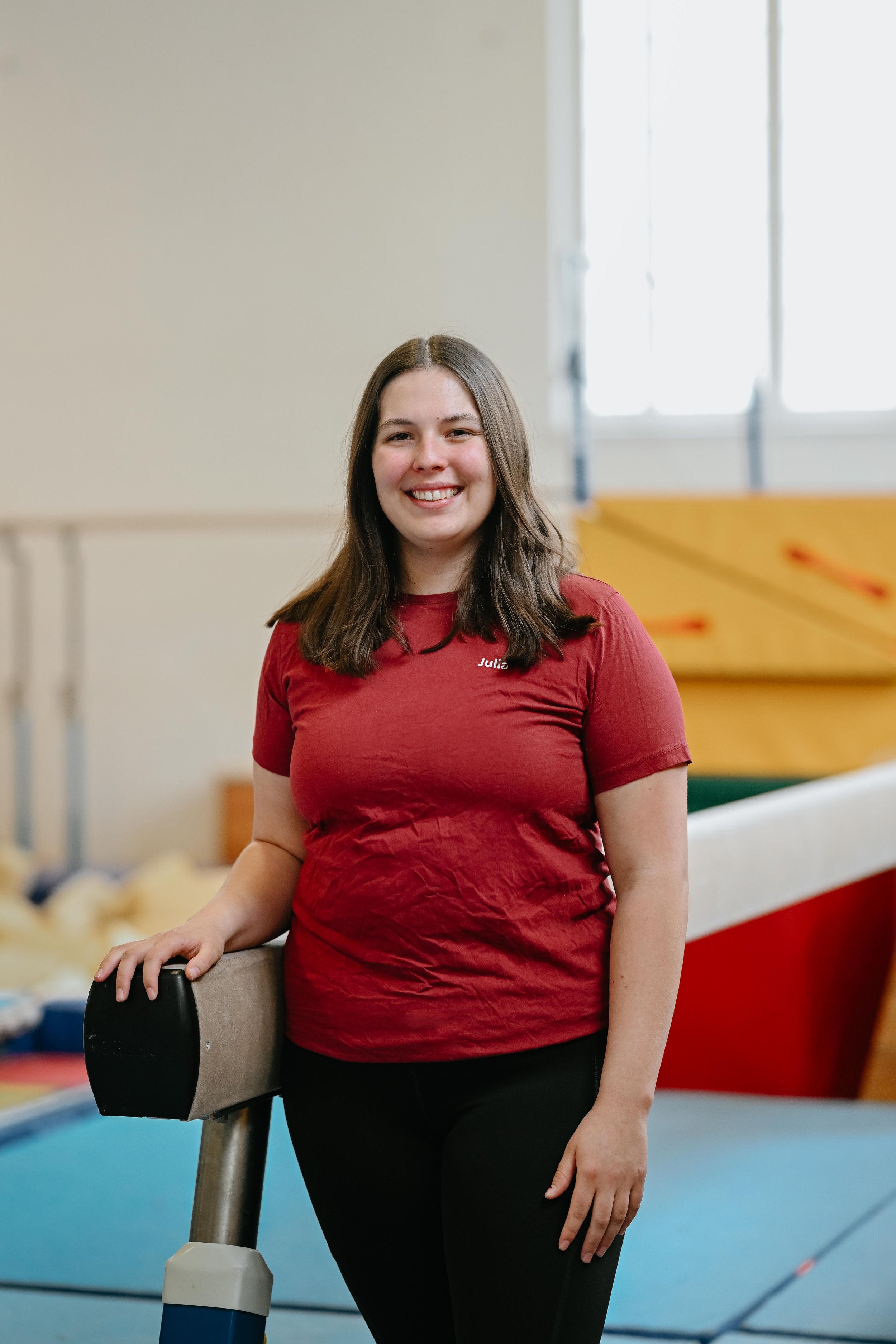 Eine junge Frau mit braunen Haaren in einem Turnanzug steht in einem Turnraum, der im Hintergrund Turngeräte enthält.