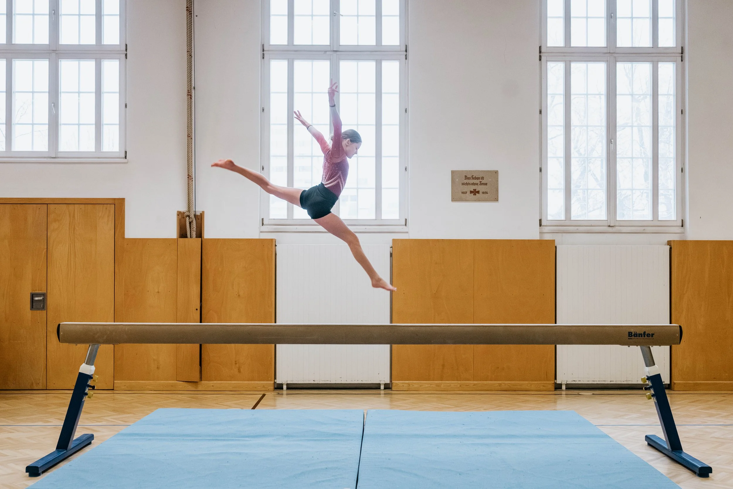 Turnende Artistin auf einem Balancebalken in einer Turnhalle, mit großen Fenstern im Hintergrund.