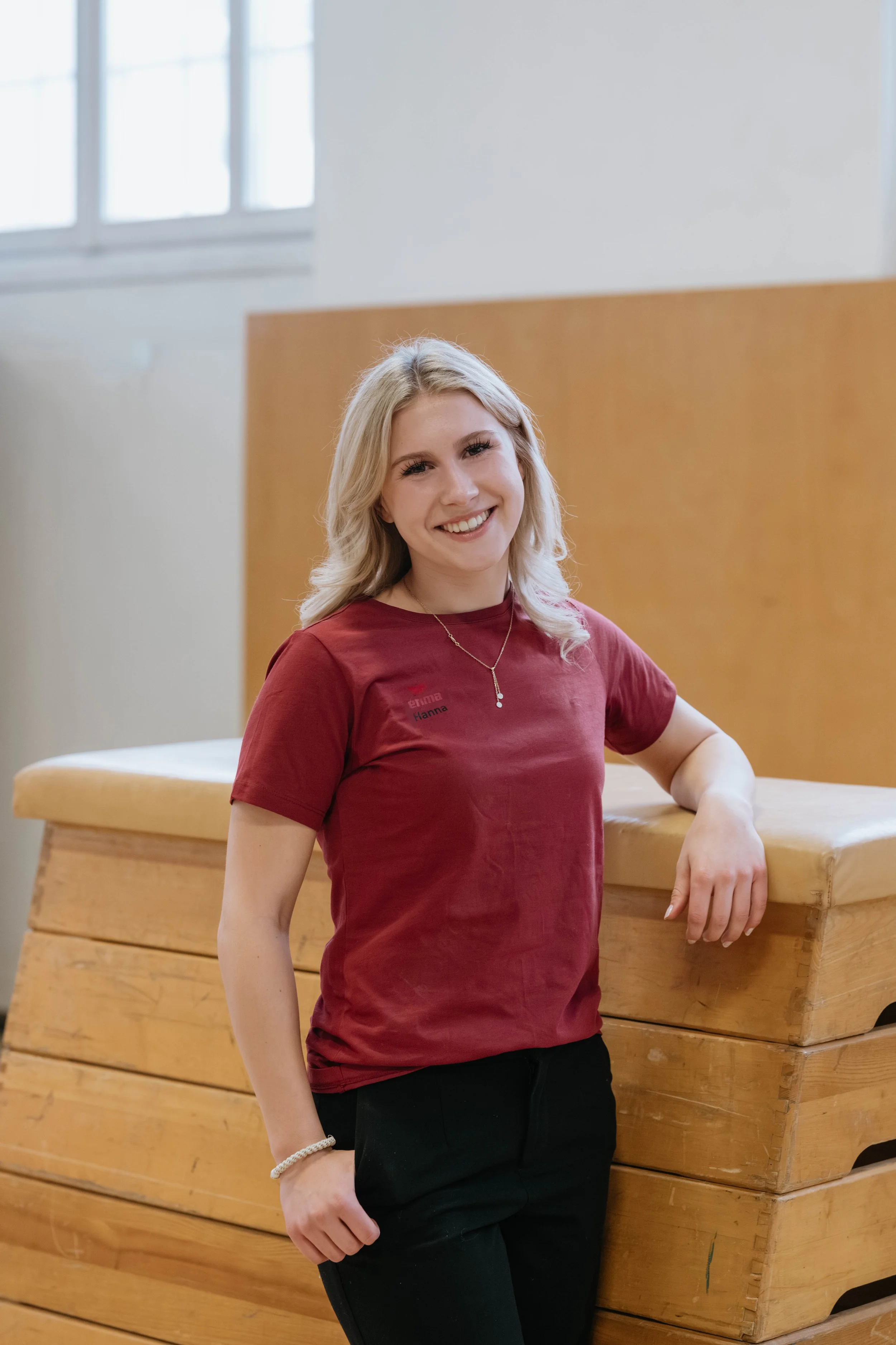Junge Frau mit blonden, gewellten Haaren trägt eine rote T-Shirt mit Namen und steht in einer Turnhalle, neben Turnmatten.