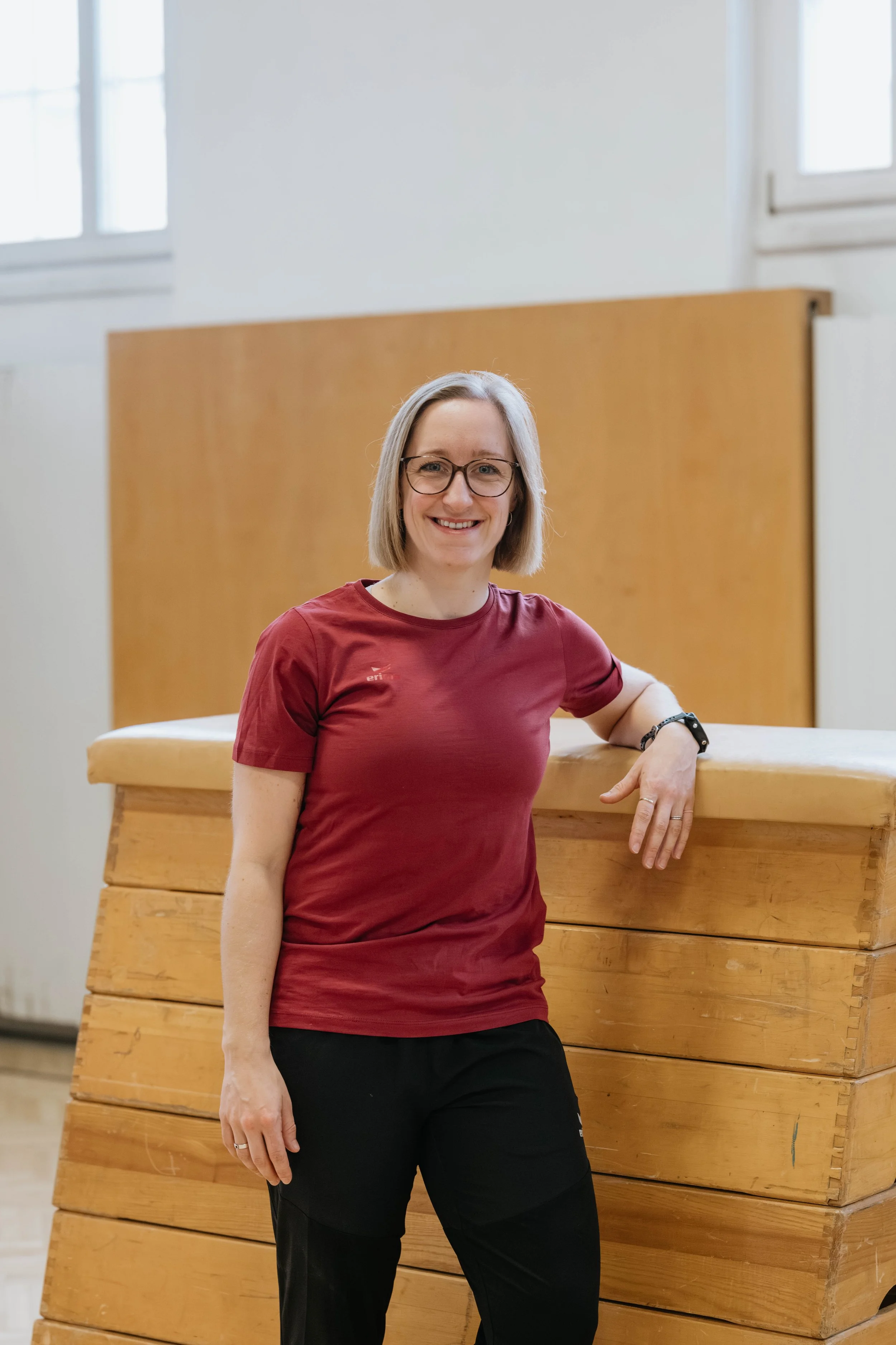 Eine lächelnde Frau mit Brille, kurzen braunen Haaren, rotem T-Shirt und schwarzen Hosen, lehnt an einem knallroten Turnen- oder Kraftgerät in einem Turnraum mit weißen Wänden und hohen Fenstern.